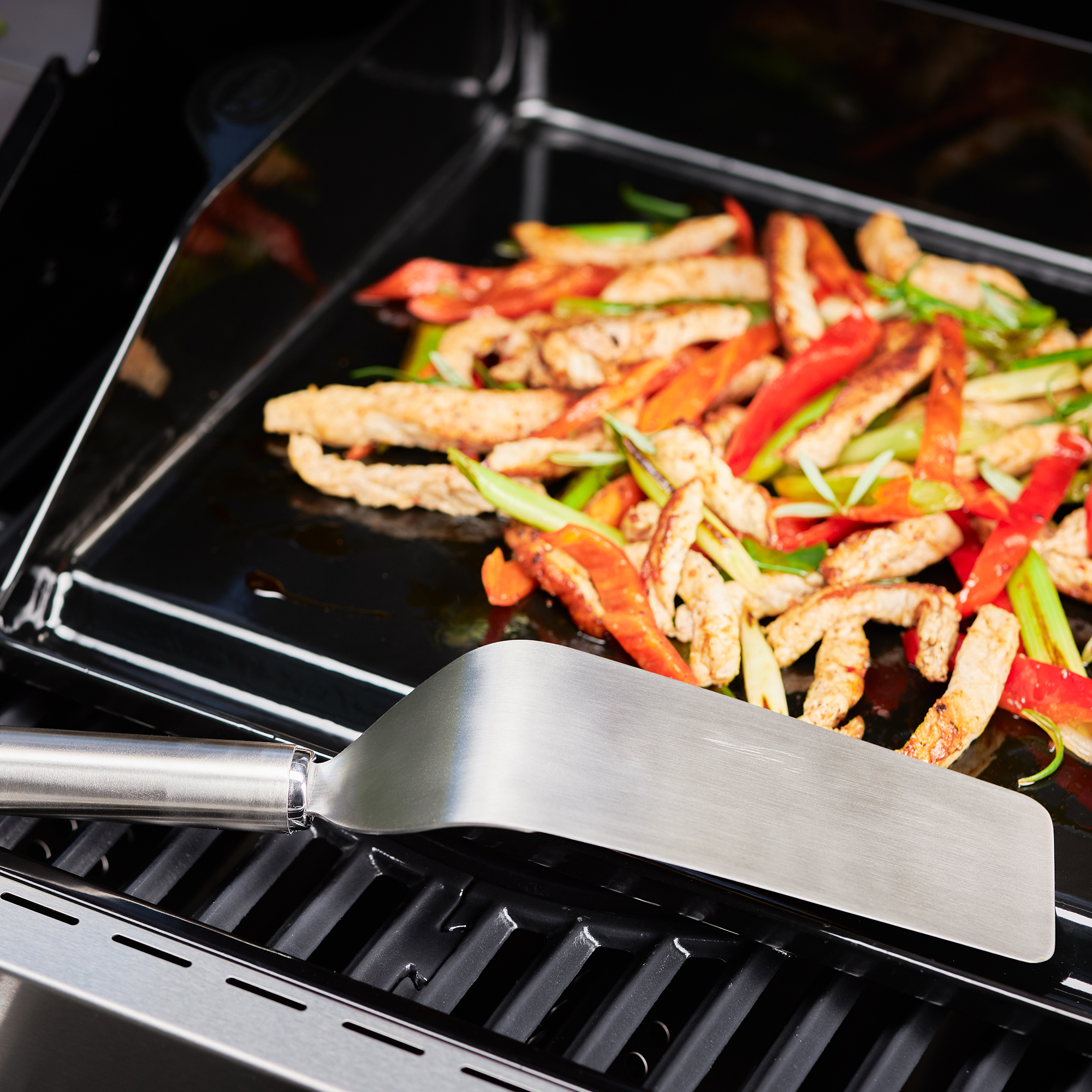 RÖSLE narrow plancha spatula with grilled vegetables Stainless steel plancha spatula, narrow, lies next to grilled vegetables on a grill plate.