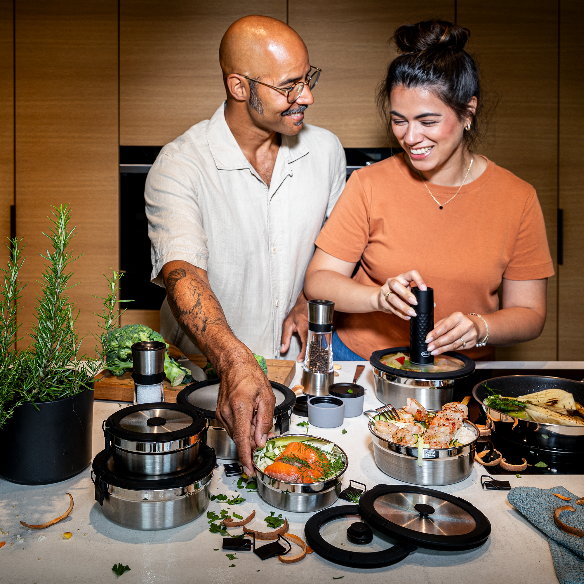 Zwei Personen in einer modernen Küche bereiten Essen mit Edelstahlbehältern vor. Eine Person hält einen Behälter mit Gemüse, die andere verwendet einen Gewürzstreuer.