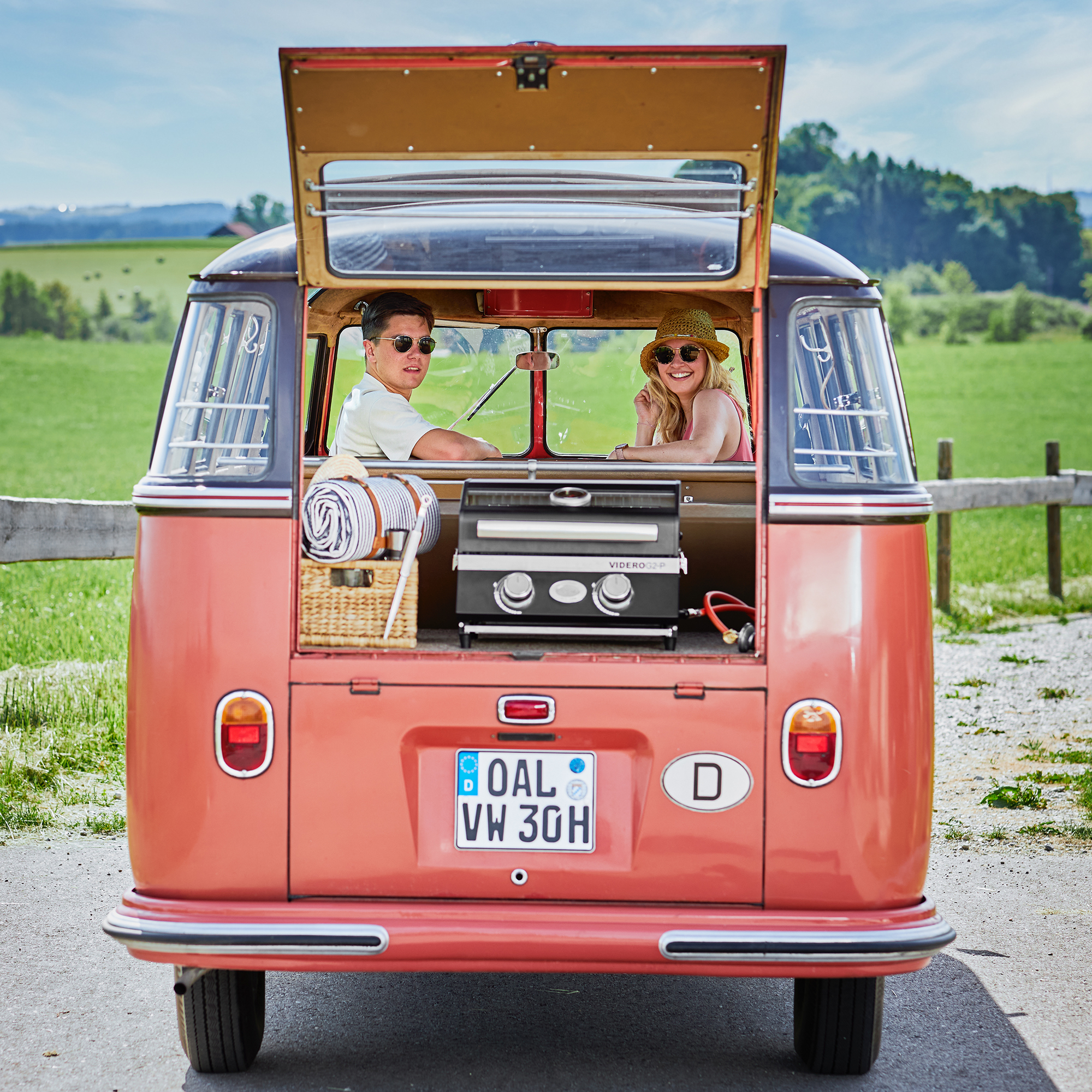 RÖSLE Gasgrill im VW-Bus Gasgrill Videro G2-P im Kofferraum eines orangefarbenen VW-Busses, mit zwei Personen im Hintergrund.