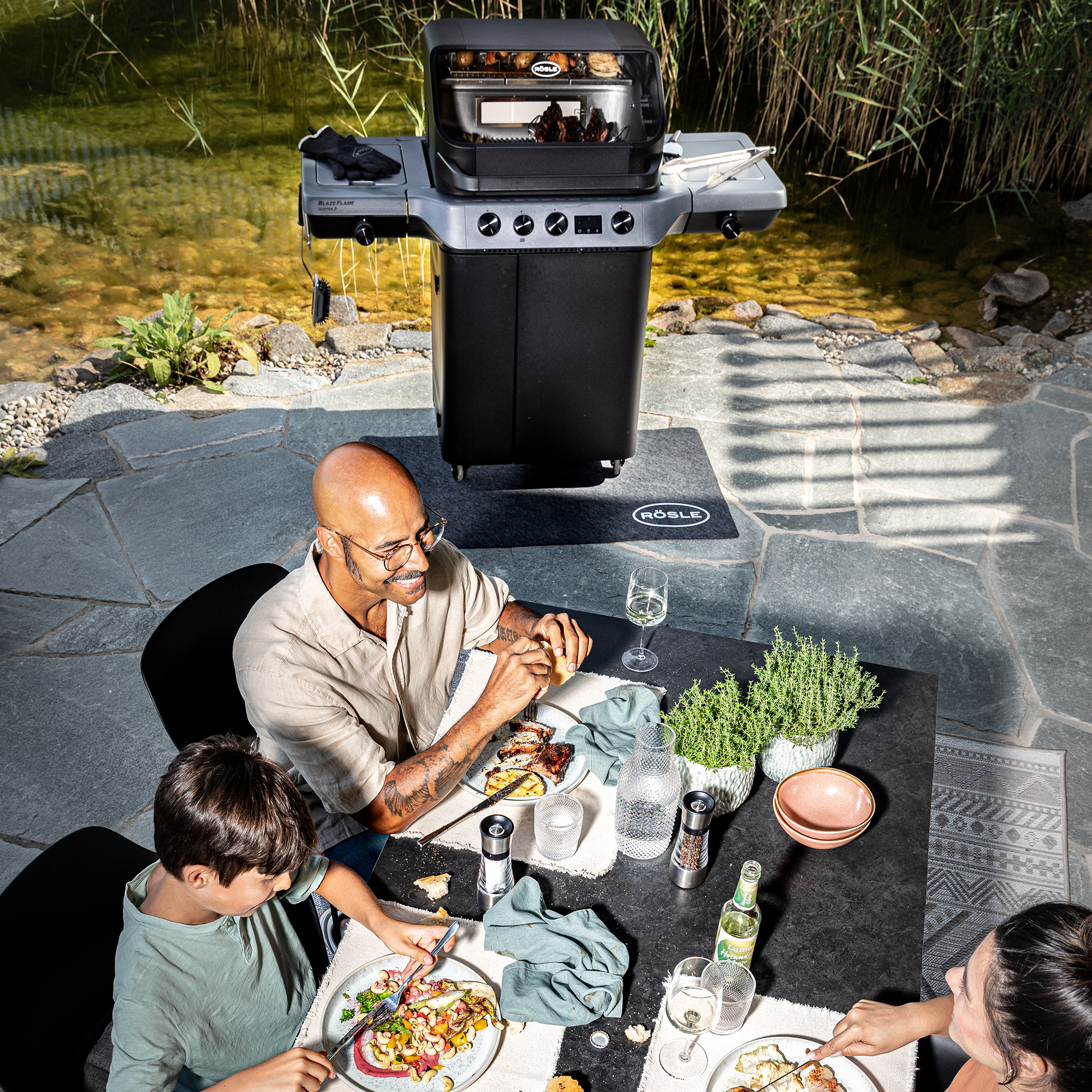 Familie genießt Essen am Tisch mit RÖSLE Grill im Hintergrund Blazeflame Master Grill im Hintergrund, Familie am Tisch mit Essen und Getränken, entspannte...
