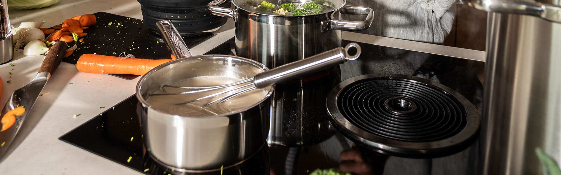 RÖSLE Stielkasserolle auf Herd Stielkasserolle aus Edelstahl auf einem Herd, umgeben von Gemüse und Kochutensilien.