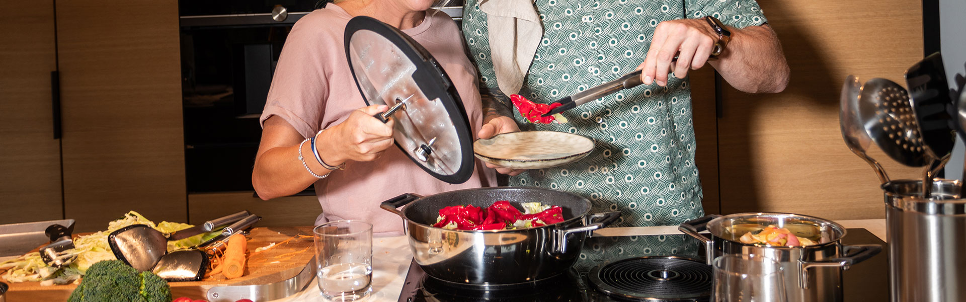 Kochszene mit Servierpfanne Zwei Personen beim Kochen in einer modernen Küche, eine Person hält einen Deckel über einer...