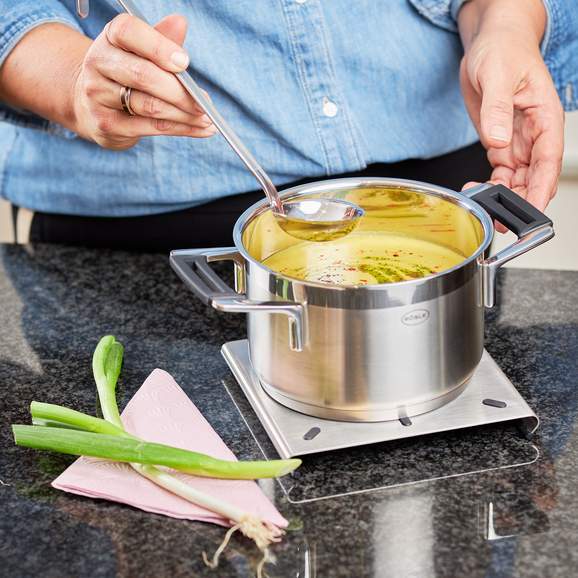 RÖSLE Edelstahl Topfuntersetzer mit Topf Topfuntersetzer aus Edelstahl, mit einem Topf auf einer Arbeitsplatte, eine Person rührt mit...