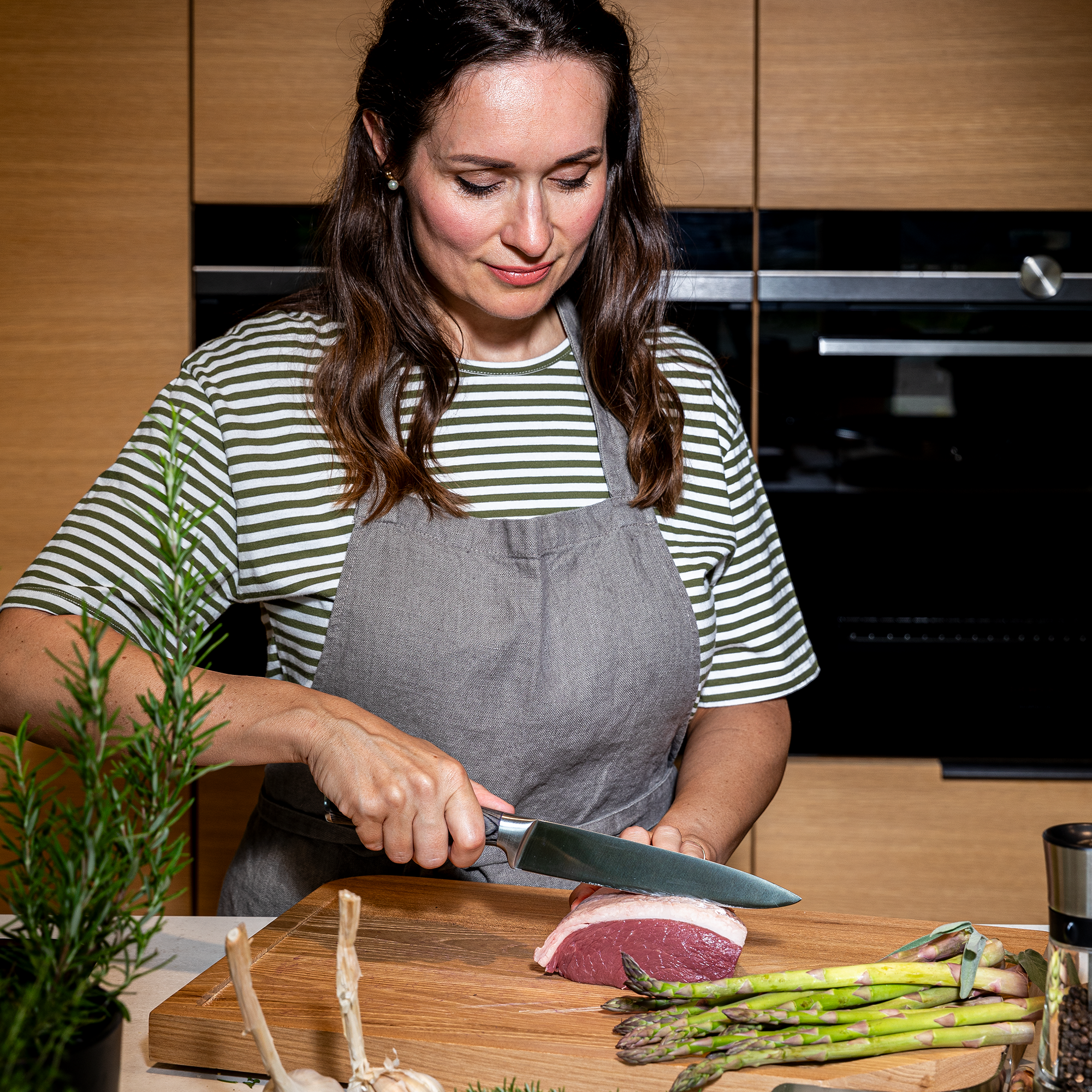Frau schneidet Fleisch mit RÖSLE Fleischmesser Fleischmesser in Wolfgrey, eine Frau schneidet Fleisch auf einem Holzbrett, umgeben von frischem...