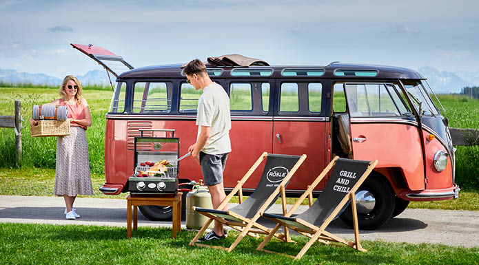 Mann am Grill und Frau mit Picknickkorb in der Hand vor VW Bully