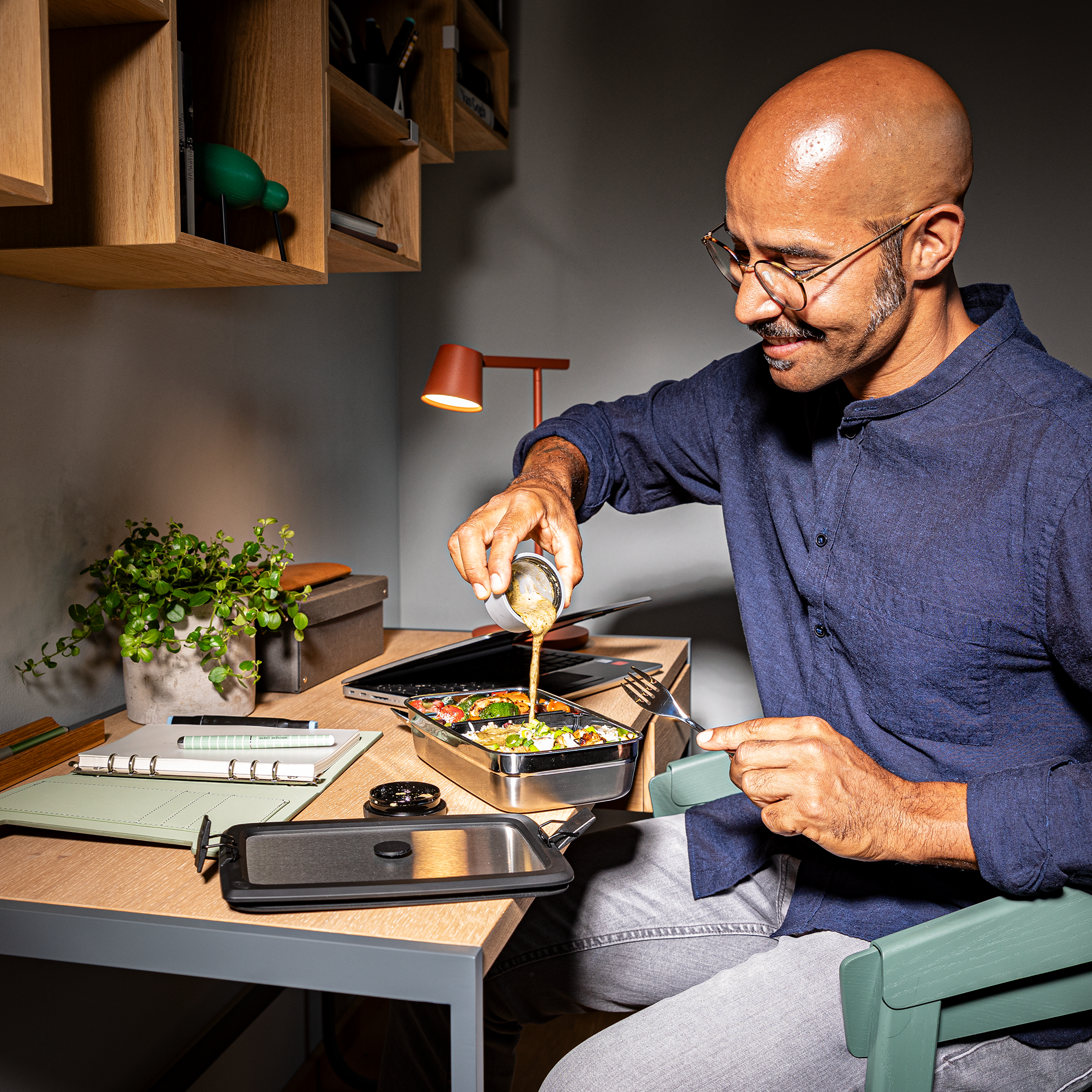 Mann in blauer Bluse, der in einer modernen Umgebung eine Dressingdose über einen Salat gießt.