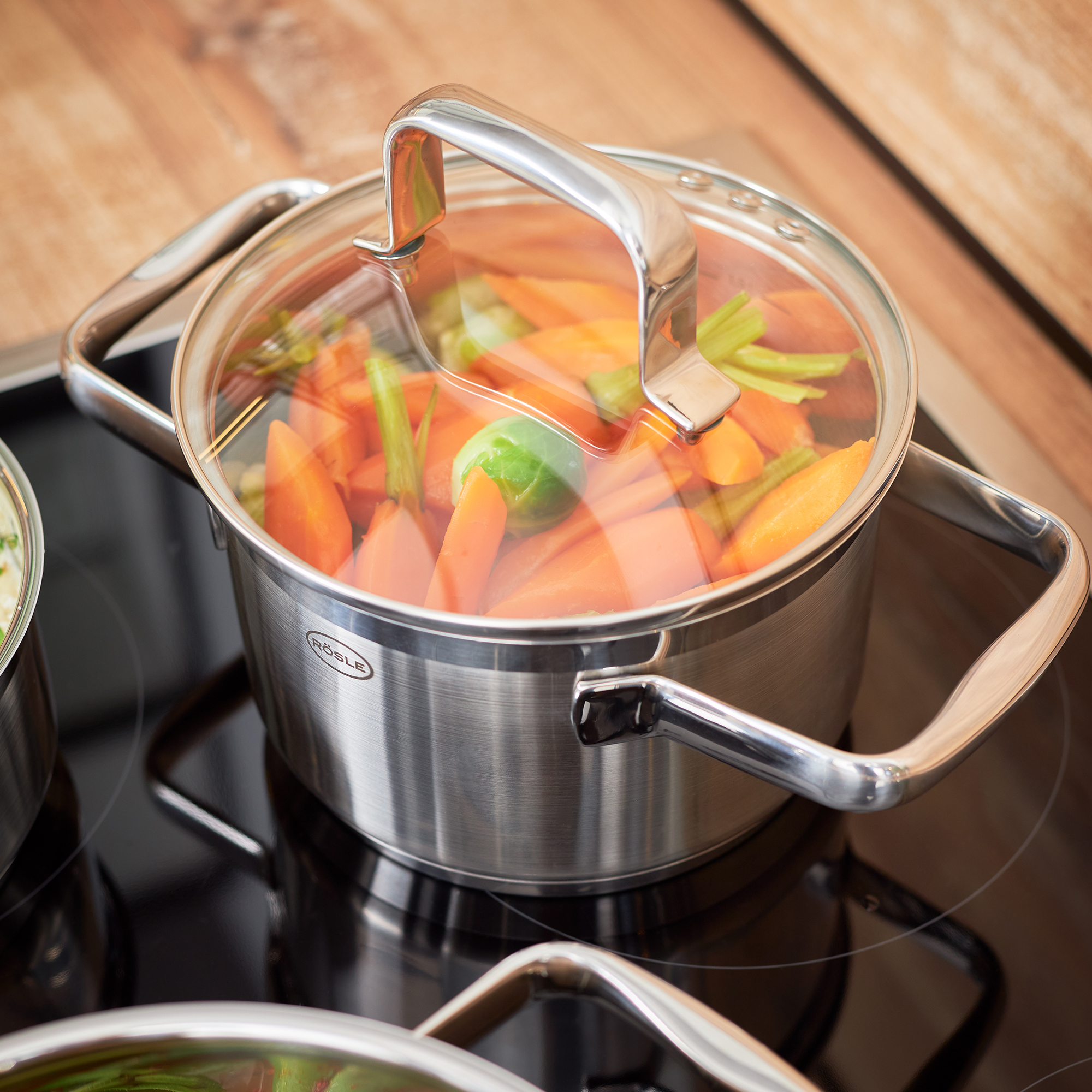 Stainless steel cooking pot with glass lid, filled with carrots and vegetables on a hob.