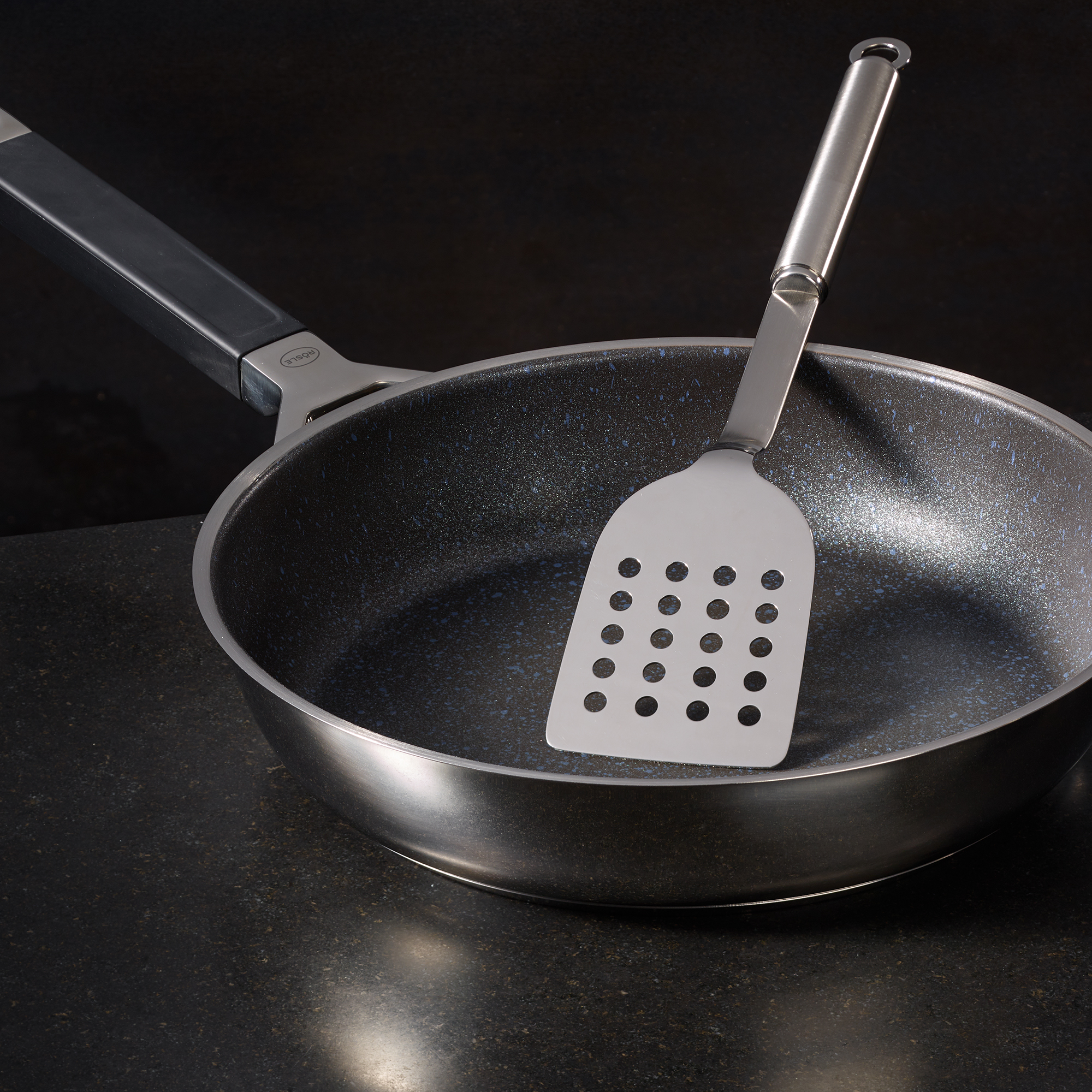 RÖSLE pan shovel in pan Stainless steel pan scoop with holes, placed in a coated pan.