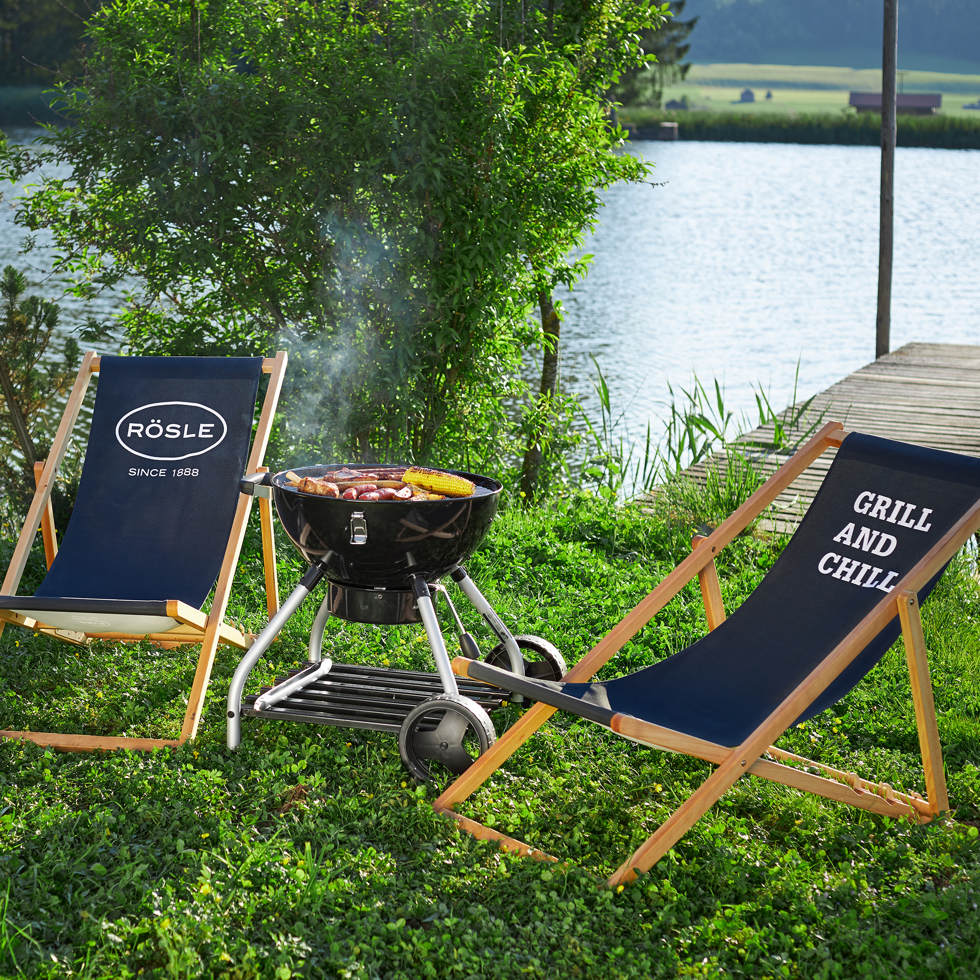 RÖSLE Holzkohlegrill im Freien Holzkohlegrill im Freien mit Grillgut, umgeben von zwei Liegestühlen mit den Aufschriften...