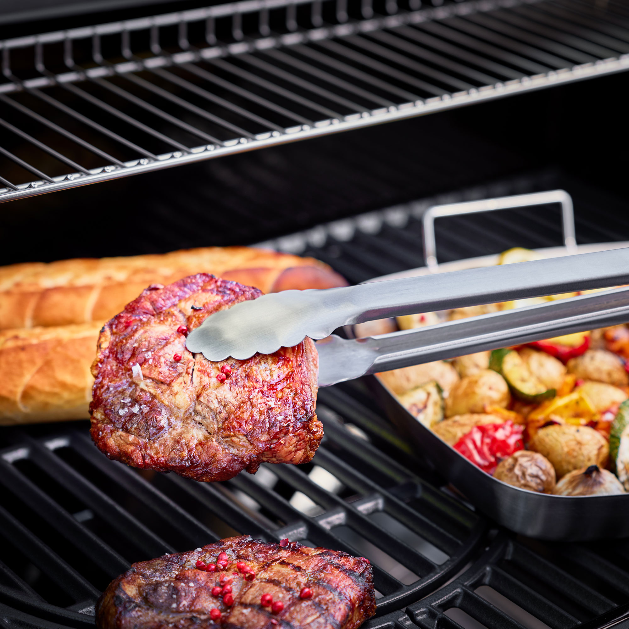 RÖSLE Grillzange mit Fleisch und Gemüse auf dem Grill Grillzange aus Edelstahl hebt ein Stück Fleisch vom Grill, im Hintergrund Gemüse und Brot...