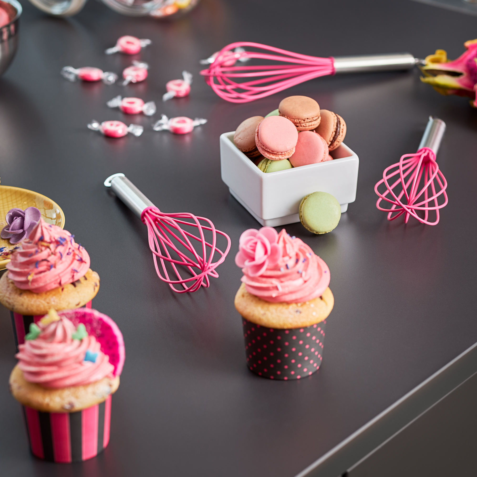 RÖSLE Pinker Schneebesen mit Cupcakes und Macarons Schneebesen in Pink, umgeben von Cupcakes mit rosa Frosting und einer Schüssel mit Macarons.