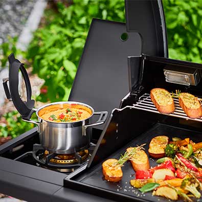 Cooking pot with soup on side burner of Magnum PRO G4 and various grilled food on main grill surface.