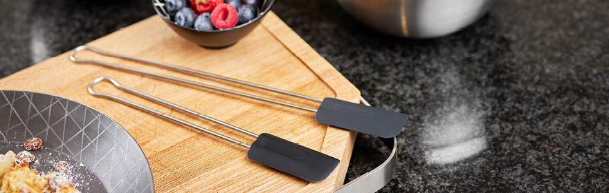 RÖSLE deegschraper op houten plank Roestvrijstalen deegschraper met zwarte siliconen kop op een houten plank, naast een kom bessen.