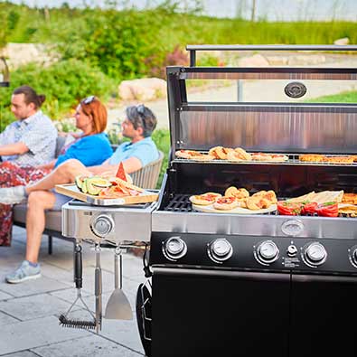 Videro G6-S with open lid and various grilled food in the foreground. In the background friends sitting on terrace
