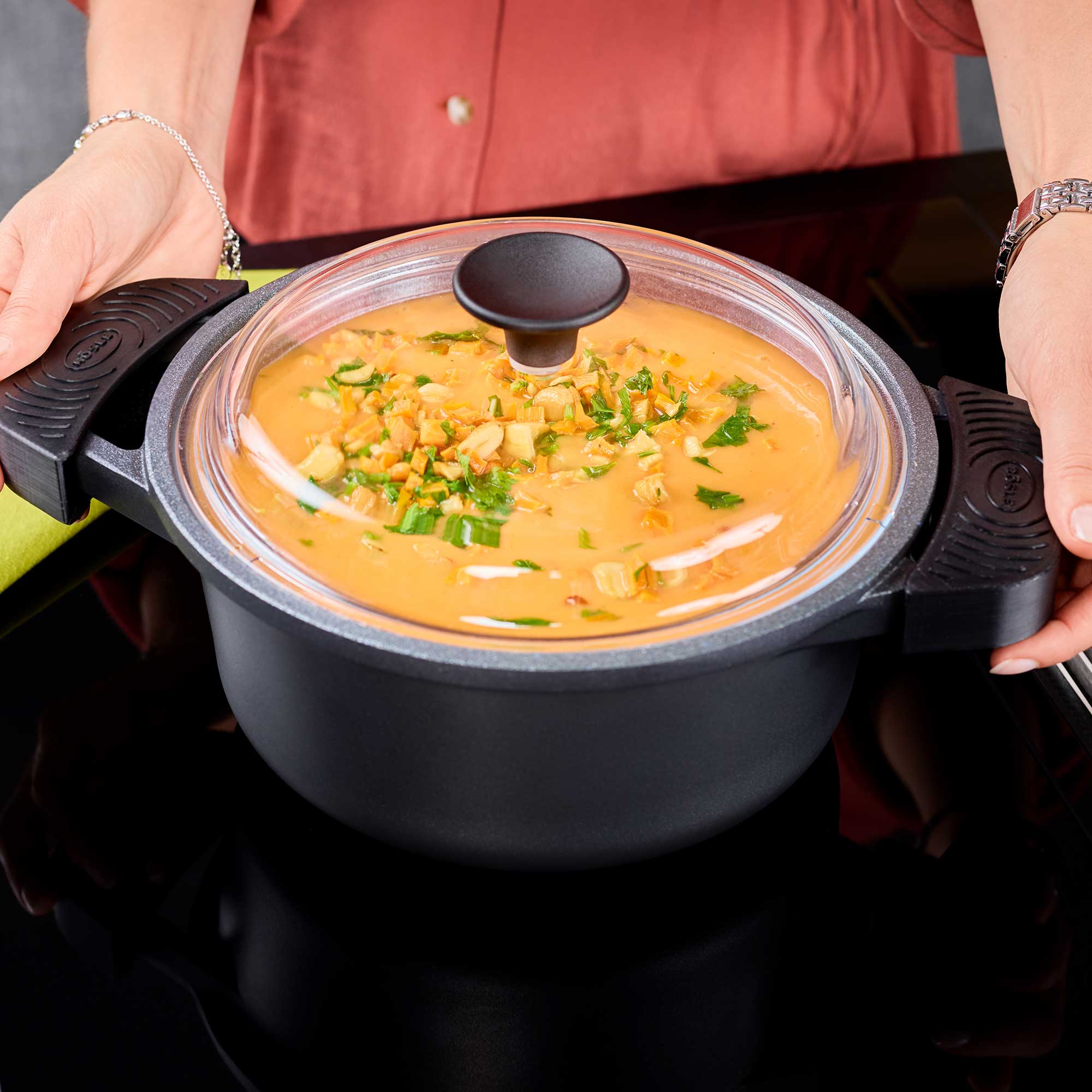 RÖSLE Kochtopf Cadini, 24 cm Kochtopf Cadini, 24 cm, schwarz mit Glasdeckel, gefüllt mit einer Suppe und Kräutern.
