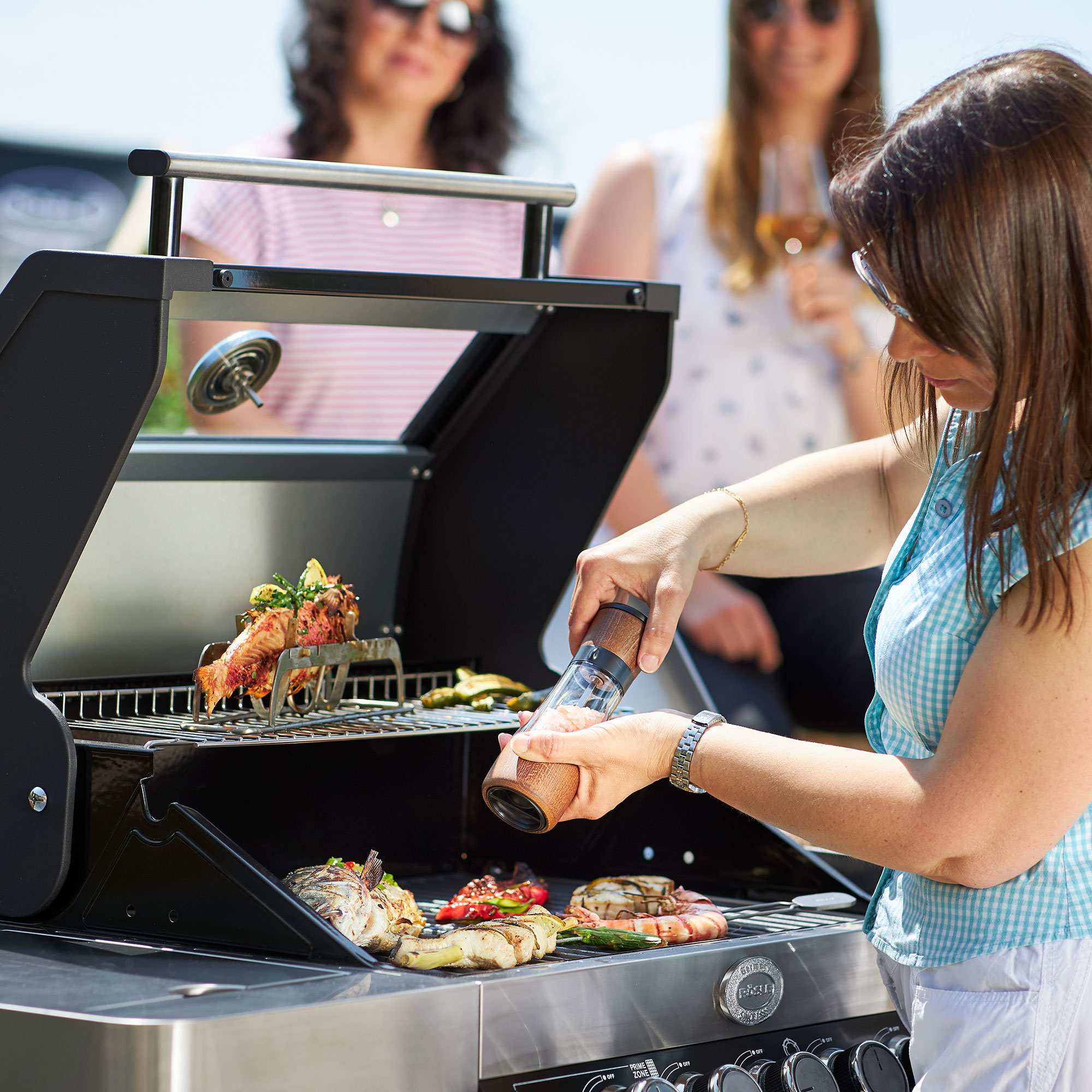 RÖSLE Fischhalter beim Grillen Fischhalter mit Grillgut, eine Frau würzt das Essen, im Hintergrund stehen Personen.