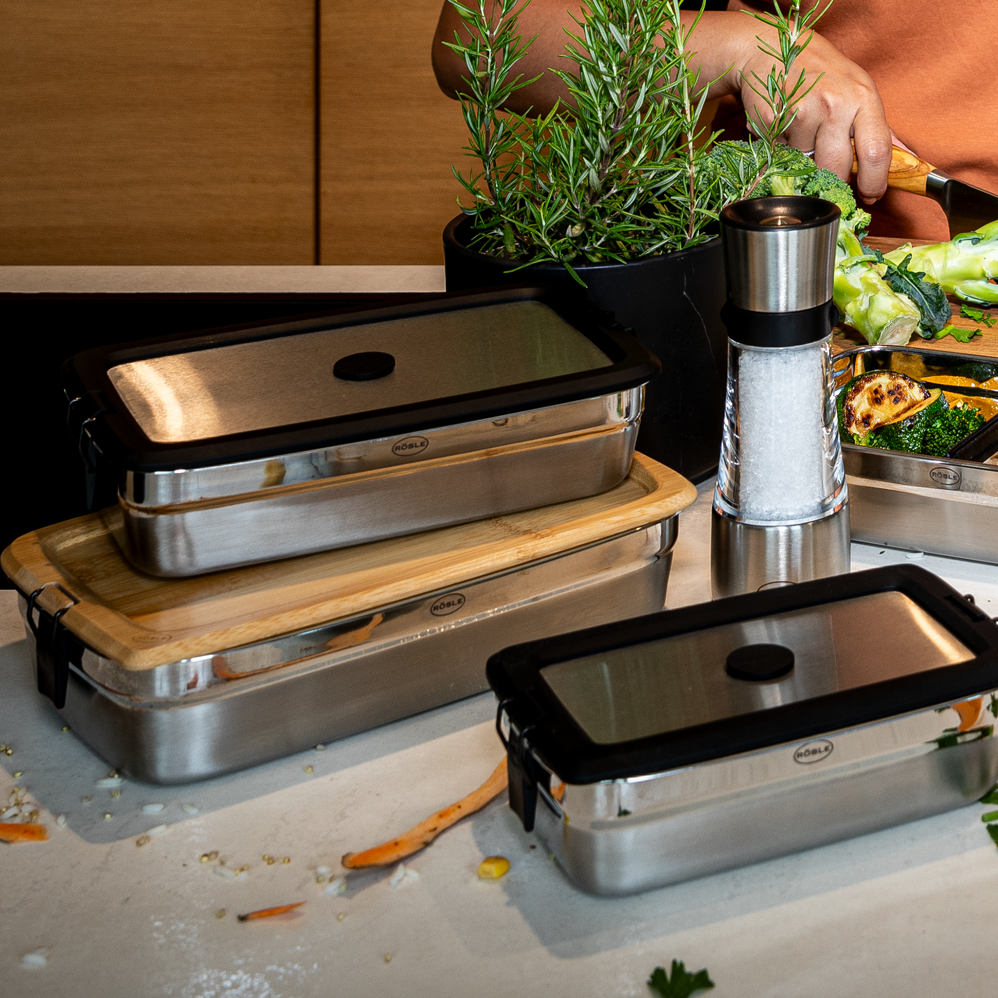 Brotzeitdeckel, eckige Edelstahlbehälter mit transparentem Deckel, auf einem Tisch mit Gemüse und Kräutern.