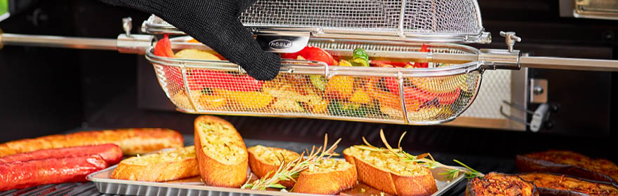 Barbecuetoebehoren met een barbecuemand die groenten vasthoudt terwijl een hand ze eruit haalt. Gegrild brood en worstjes op de achtergrond.