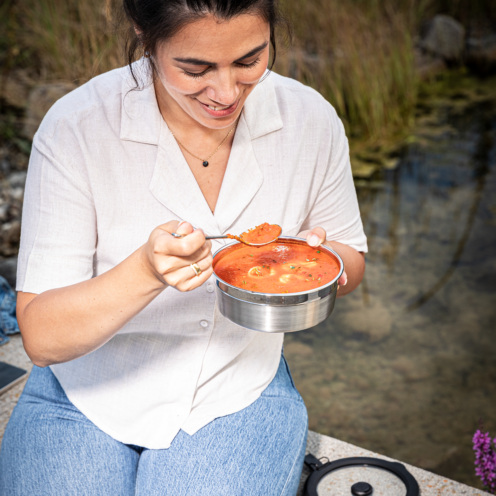 Frau mit einem Flexx2go 0,8l Behälter, der mit Suppe gefüllt ist, sitzt am Wasser und genießt das Essen.