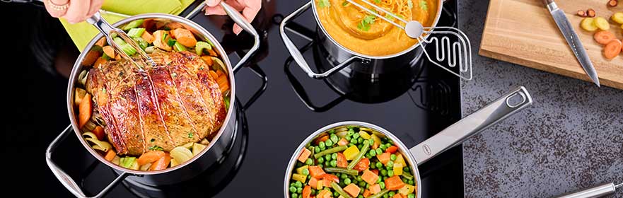 RÖSLE pannenset op fornuis Pannenset met verschillende potten gevuld met groenten en een braadstuk op een kookplaat.