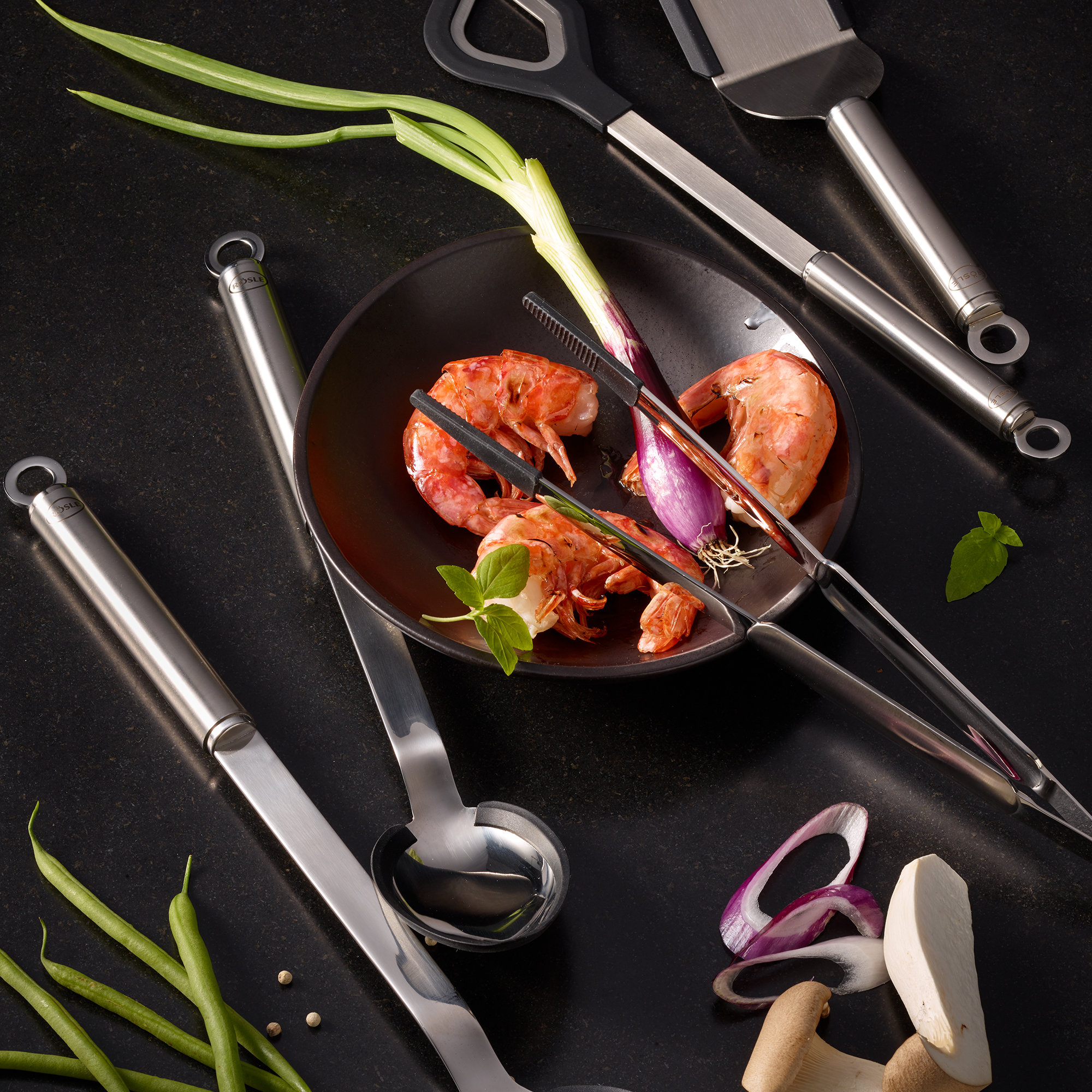 RÖSLE round-handled watering spoon and kitchen utensils Round-handled silicone pouring spoon surrounded by various kitchen utensils and fresh ingredients...