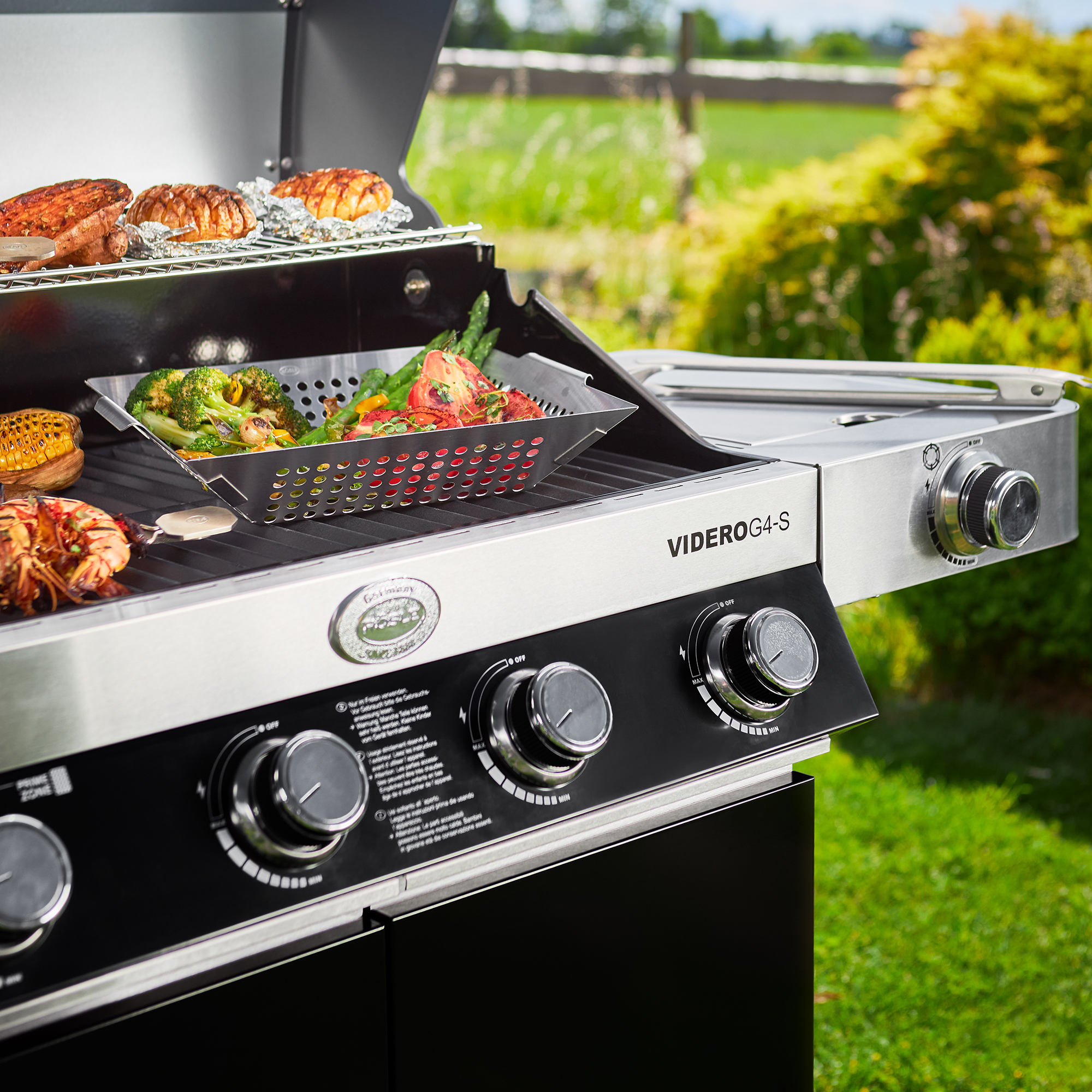 RÖSLE Videro G+S Grill mit Gemüsepfanne Videro G+S Grill mit Gemüsepfanne, Edelstahl, auf einem Gartenplatz mit Grillgut.