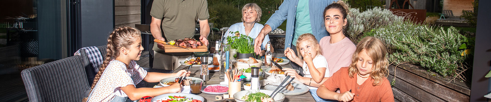 Geschichte Familie sitzt zusammen mit Kindern am Tisch und Mann serviert gegrilltes Essen.