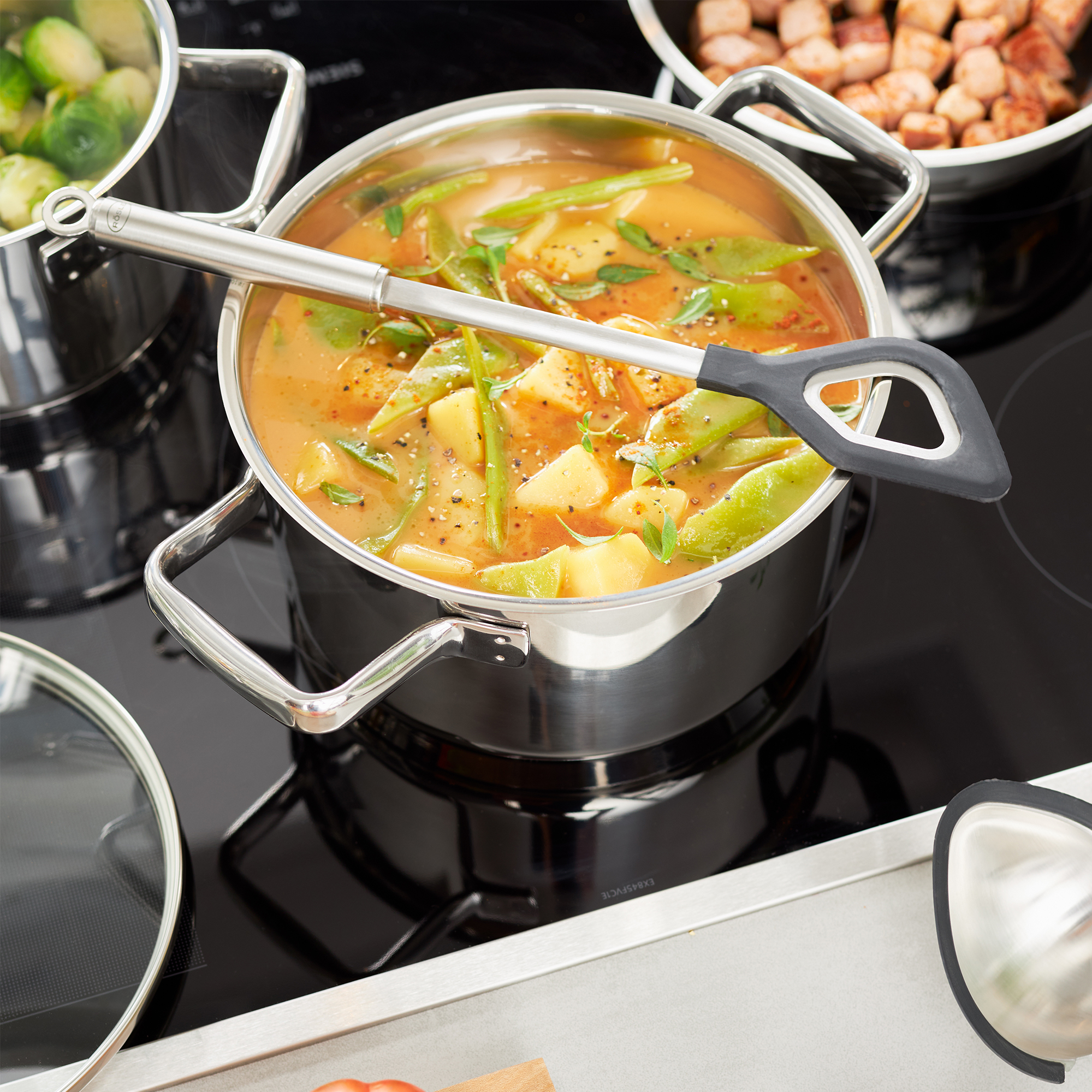 RÖSLE cooking spoons in use Round-handled silicone cooking spoon, in a pot of vegetable stock, on a cooker.