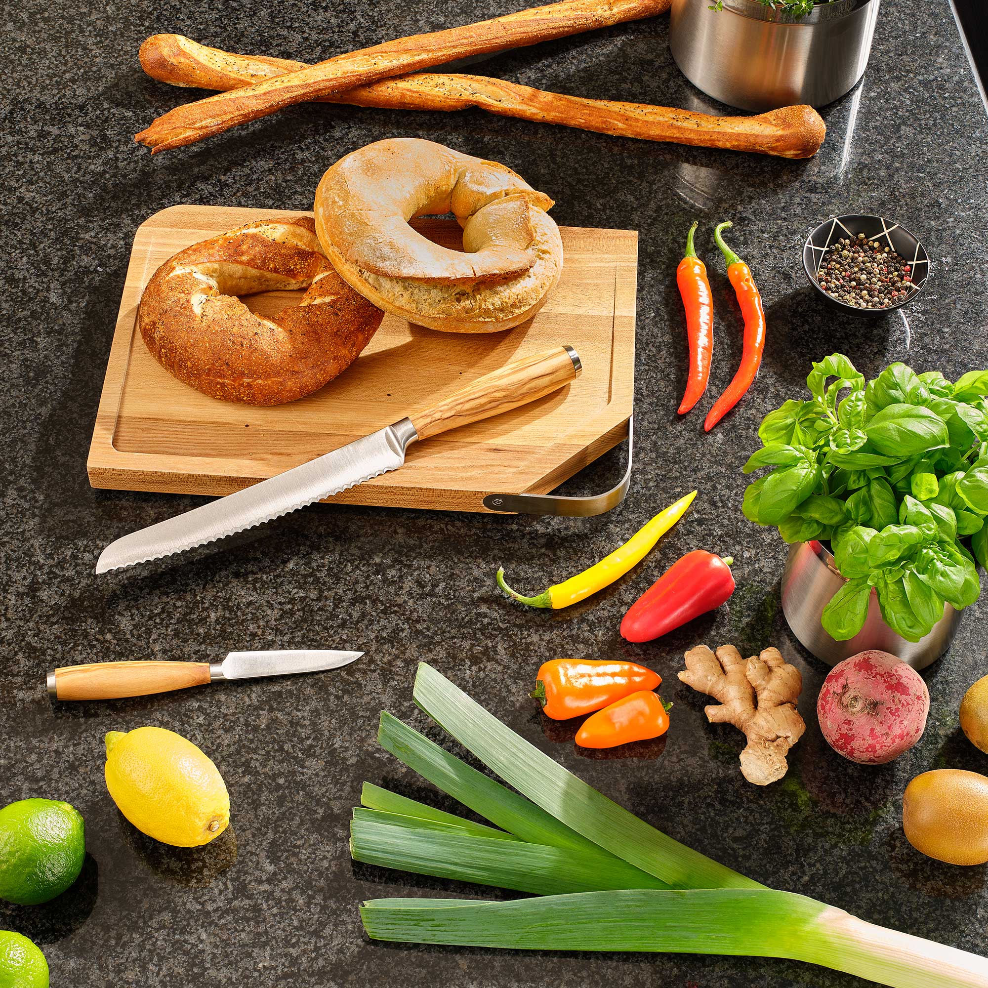 RÖSLE Brotmesser auf Holzbrett mit Gemüse Brotmesser, 22 cm, mit Holzgriff, auf einem Holzbrett neben frischem Gemüse und Backwaren.