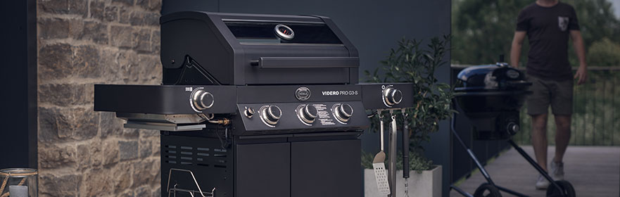 Gasbarbecue met verschillende branders, zwarte behuizing, buiten geplaatst, een andere barbecue op de achtergrond.