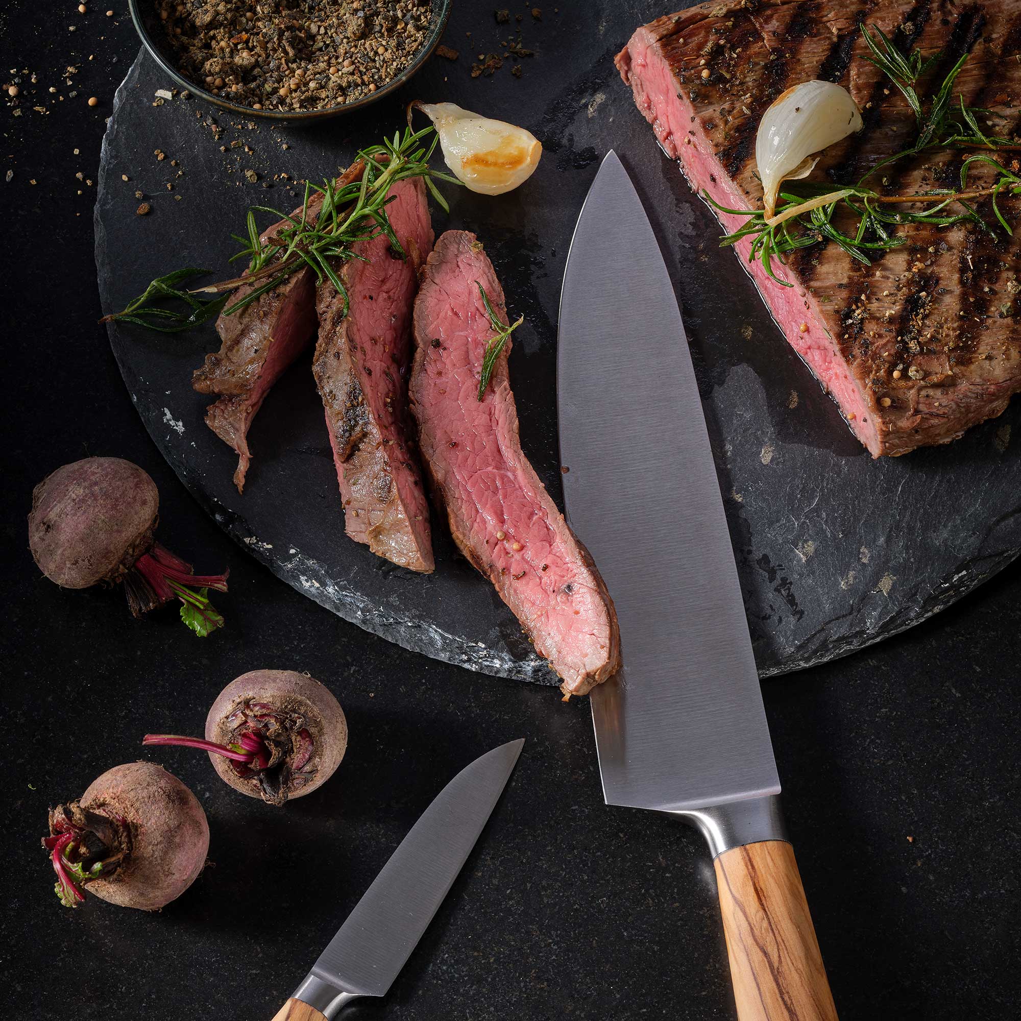 RÖSLE Fleischmesser und Steak auf schwarzem Untergrund Fleischmesser 19 cm mit Holzgriff, neben geschnittenem Steak und Gemüse auf einem schwarzen...