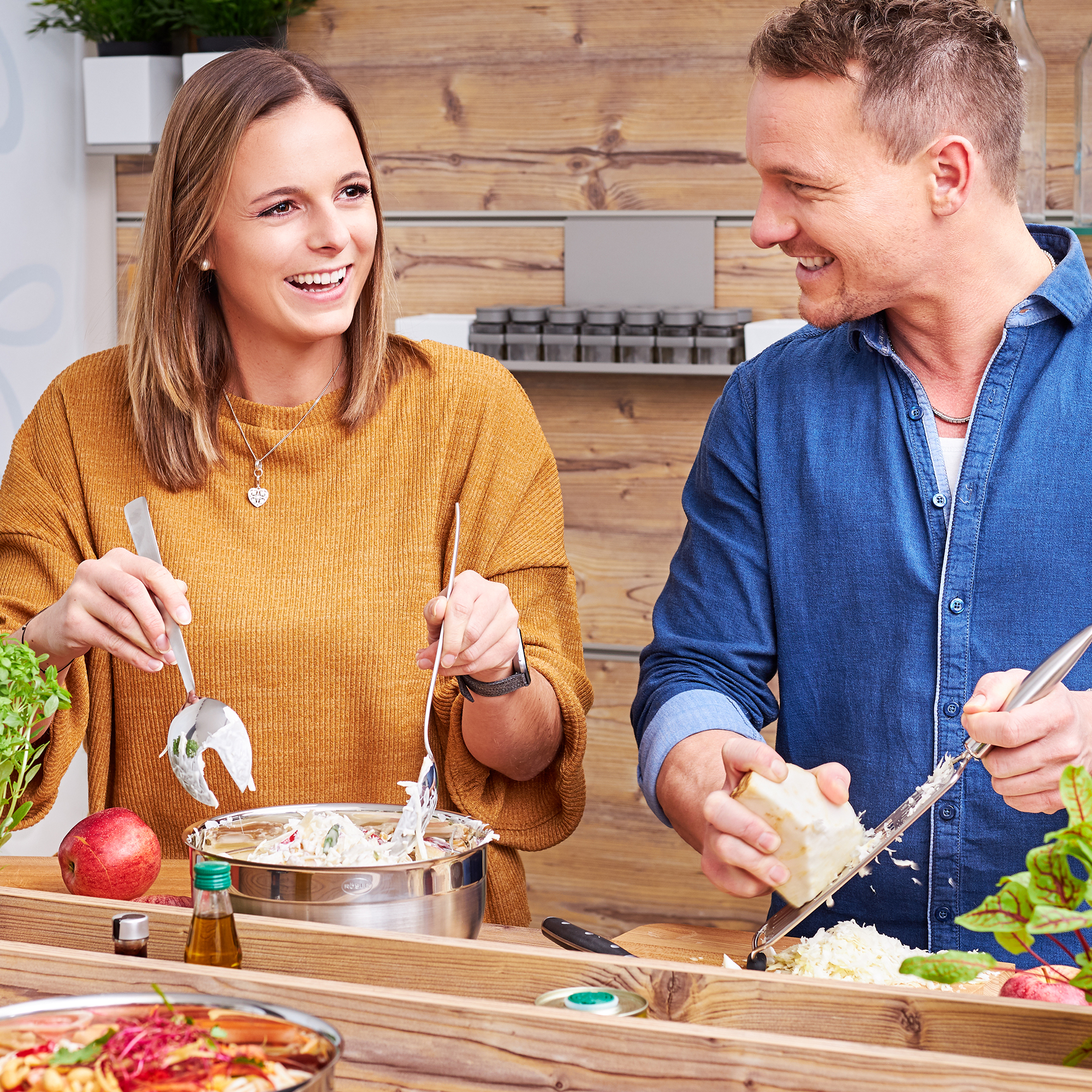 Two people preparing salad Two people in a modern kitchen, a woman with salad servers, a man with a cheese grater, both...