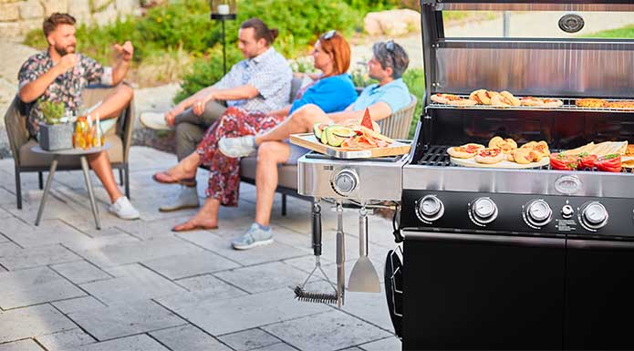 Videro G6-S with open lid and various grilled food in the foreground. In the background friends sitting on terrace
