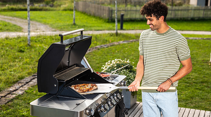 Pizza Man stands at the grill with a pizza peel and picks up a finished pizza from the pizza stone