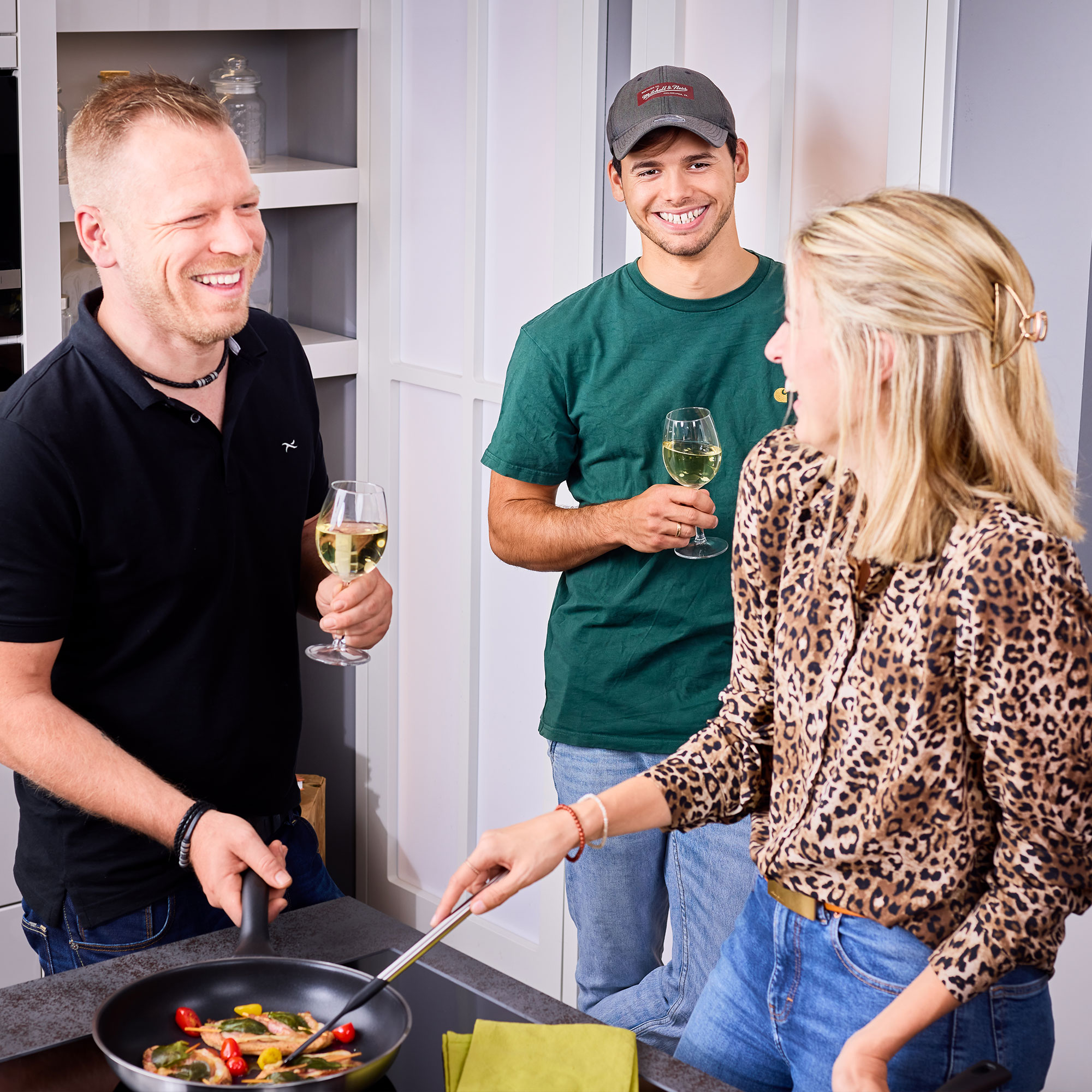 Drei Personen in einer modernen Küche, eine Person kocht in einer Pfanne, während die anderen mit Getränken anstoßen.
