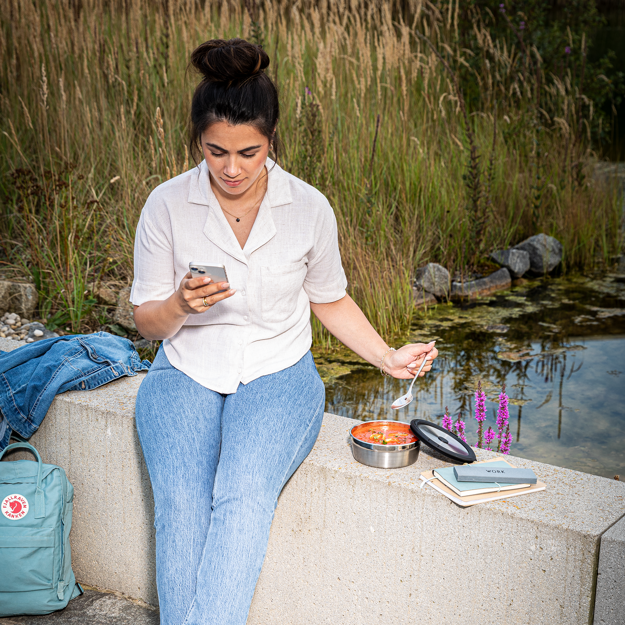 Frau mit dunklen Haaren in einem weißen Hemd sitzt am Wasser und isst aus einer Flexx2go Dose, während sie auf ihr Handy schaut.