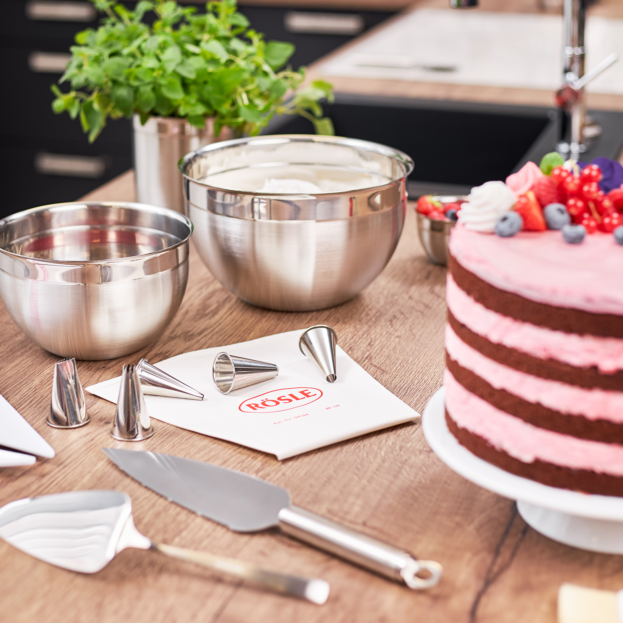 Edelstahl-Schüsseln und Backutensilien von RÖSLE Edelstahl-Schüsseln, verschiedene Größen, mit Backutensilien und einer Torte auf einem Holztisch.