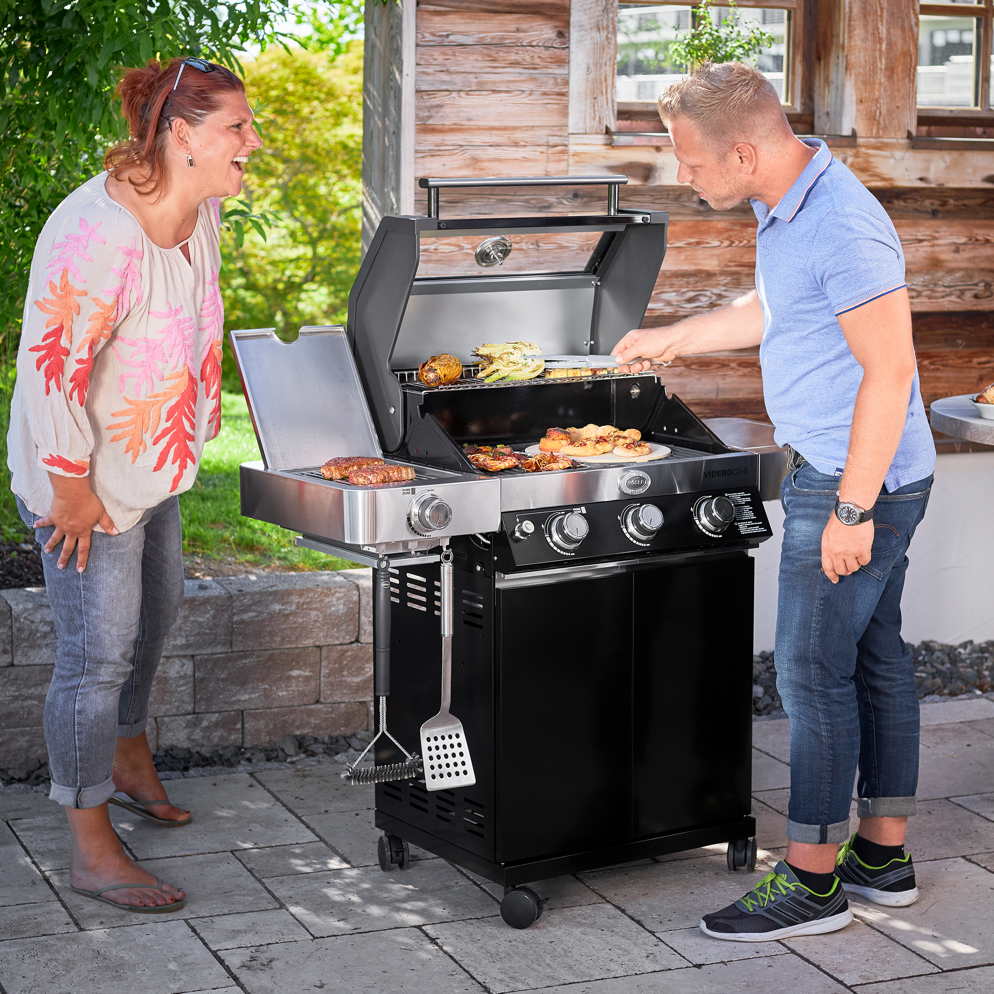 RÖSLE gas barbecue in action Gas barbecue with open lid, two people barbecuing, a woman laughing, a man holding a barbecue...