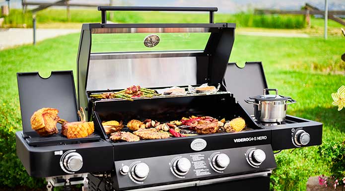 Various grilled food on prime zone and main grill surface of the gas grill. On side burner is a closed cooking pot.