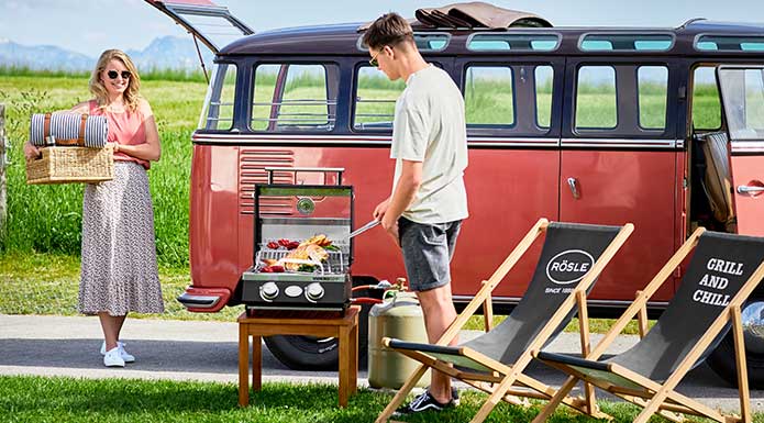 Frau räumt Picknickkorb aus VW Bully während Mann am Videro G2-P grillt.