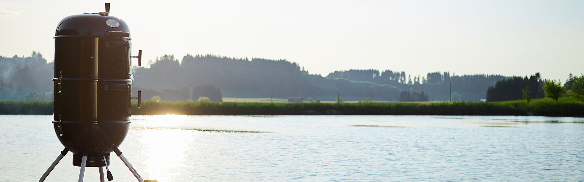 RÖSLE Smoker am See Smoker in Schwarz am Ufer eines ruhigen Sees mit grüner Landschaft im Hintergrund