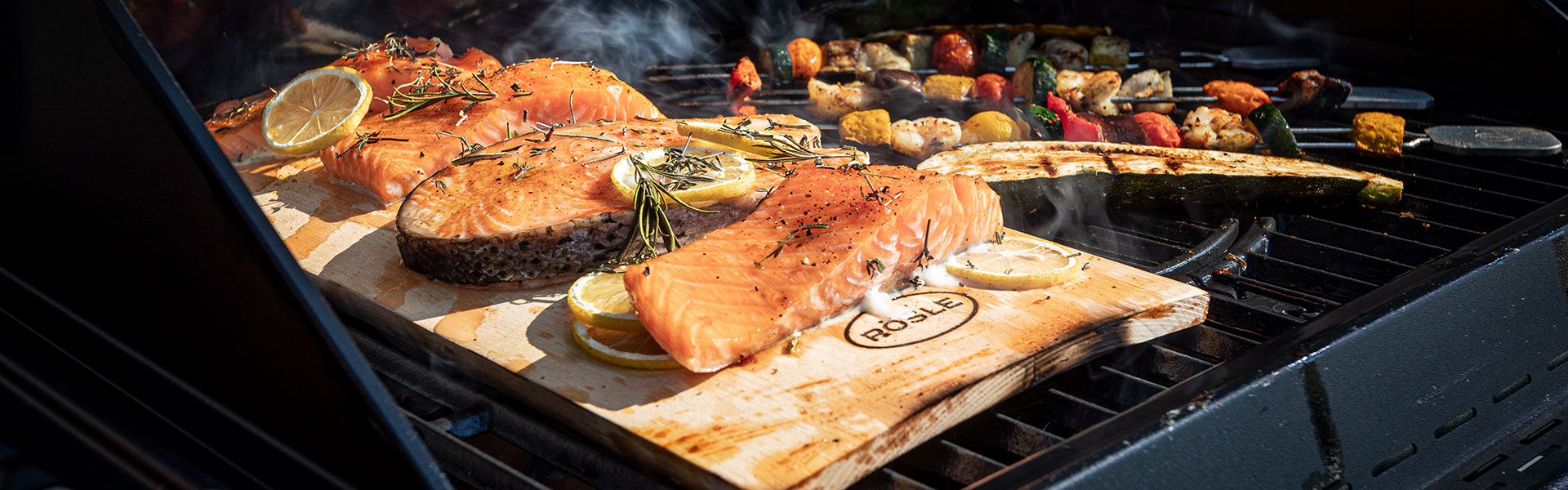 Grillwerkzeuge mit Lachs und Gemüse Grillwerkzeuge mit Lachsfilets und Gemüse auf dem Grill, umgeben von Rauch und Kräutern.