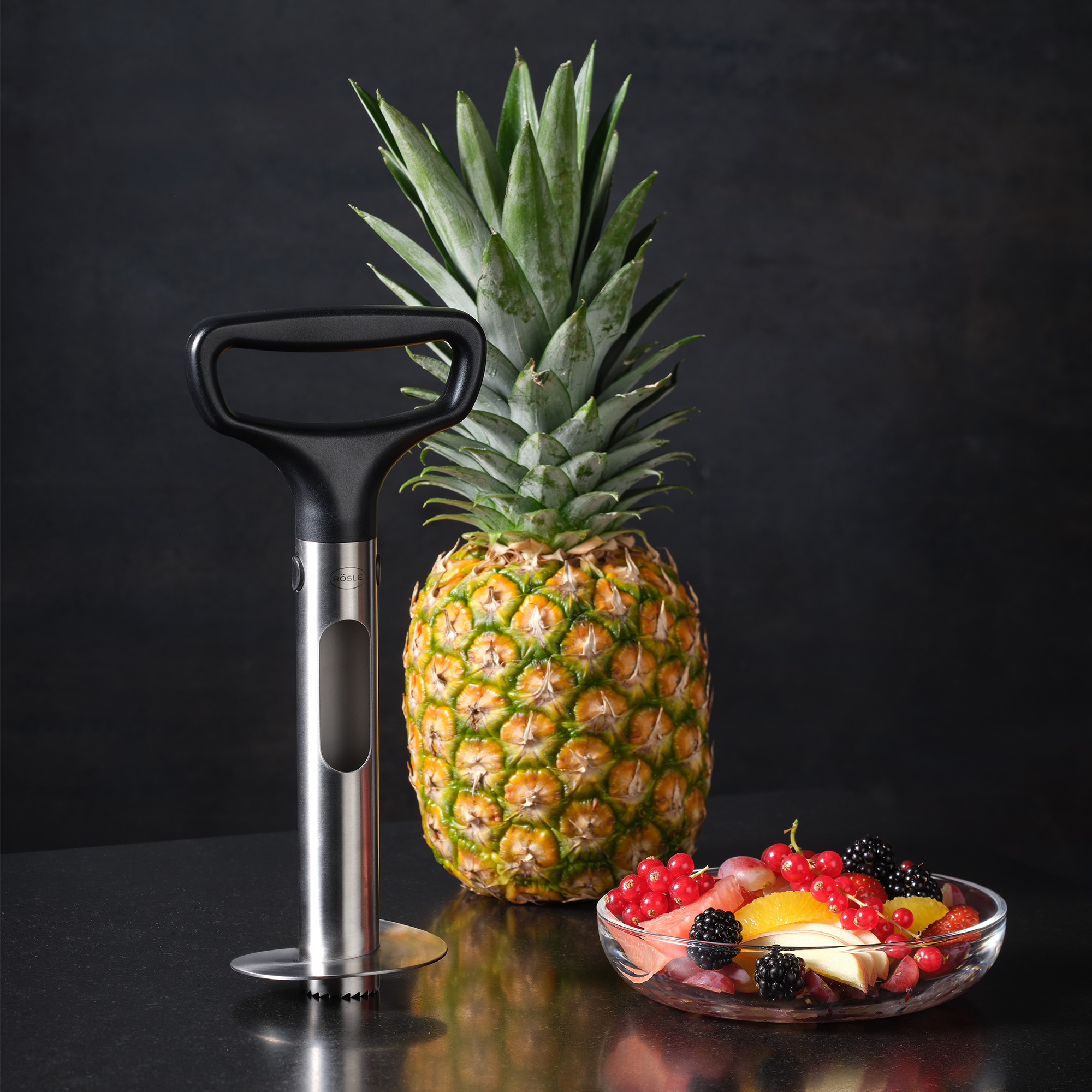 RÖSLE pineapple slicer and pineapple Stainless steel pineapple slicer with black handle, next to a pineapple and a bowl of mixed...