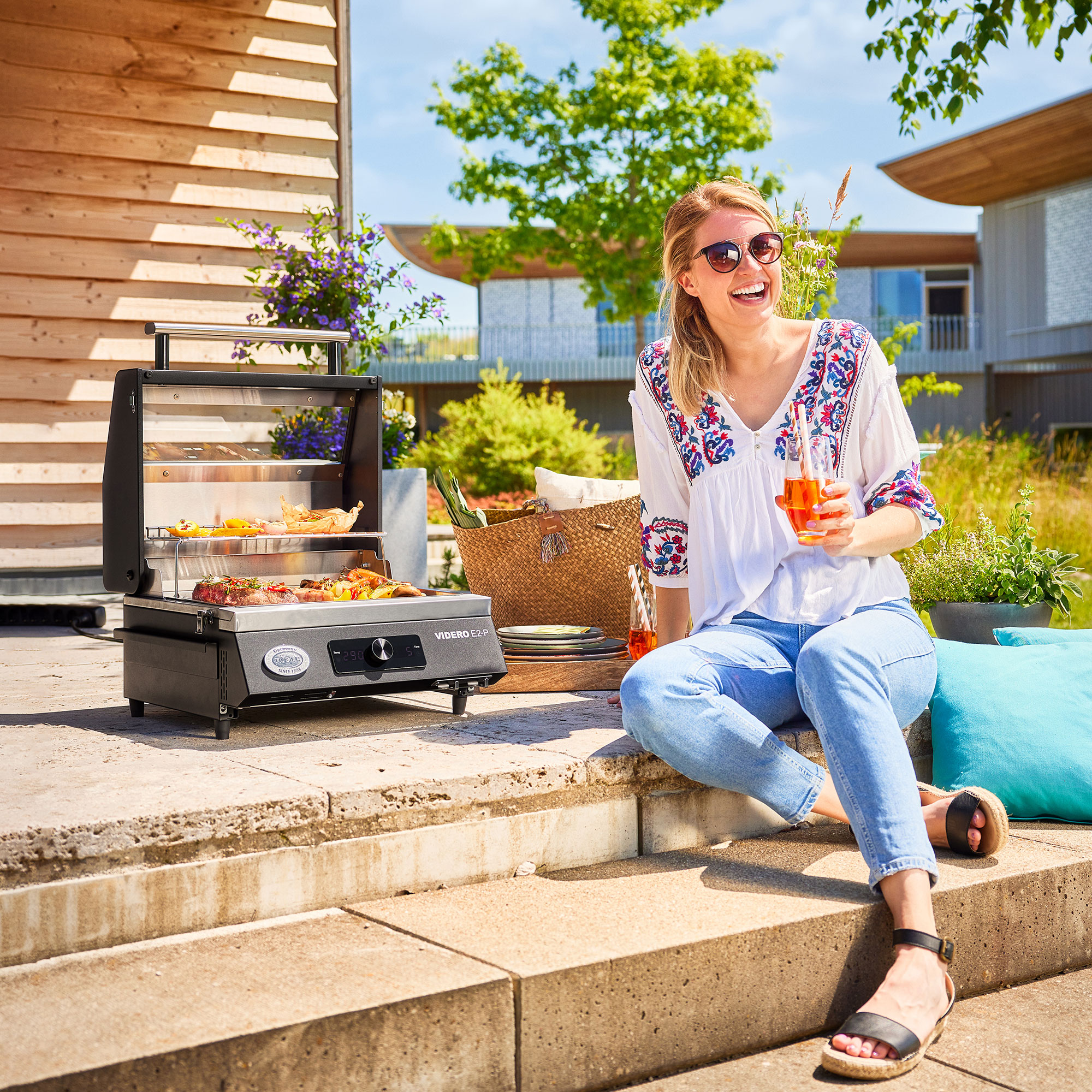 RÖSLE Elektrogrill Videro E2-P auf Terrasse Elektrogrill Videro E2-P auf einer Terrasse, eine lächelnde Frau mit Sonnenbrille hält ein...
