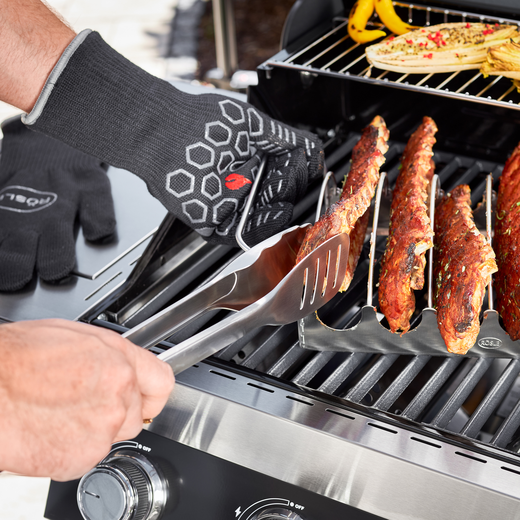 RÖSLE Premium barbecue gloves for barbecuing Premium barbecue gloves in black, one hand grasping grilled meat with barbecue tongs.