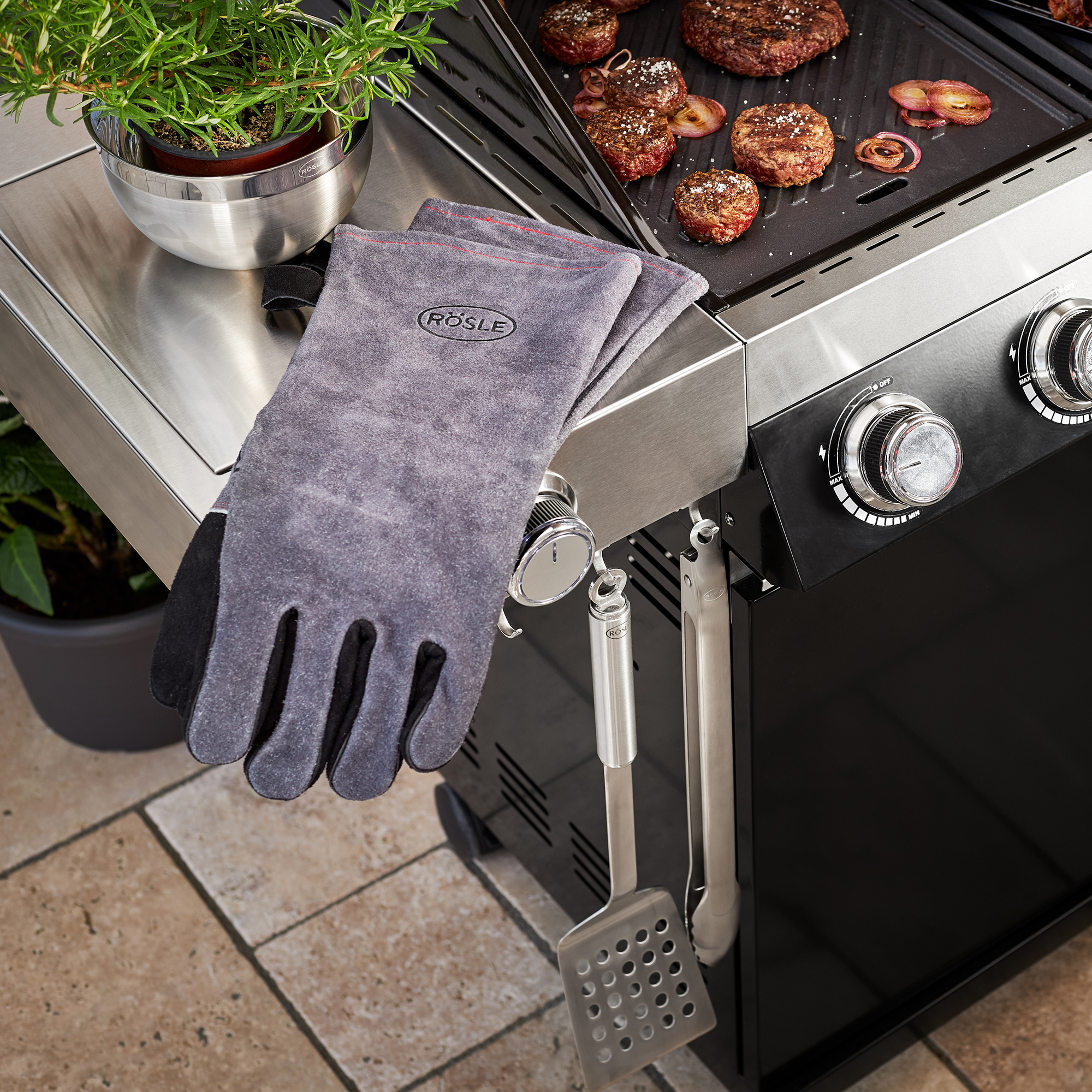 RÖSLE Grillhandschuhe Grillhandschuhe aus grauem Material, neben einem Grill mit Burgern und Zwiebeln.