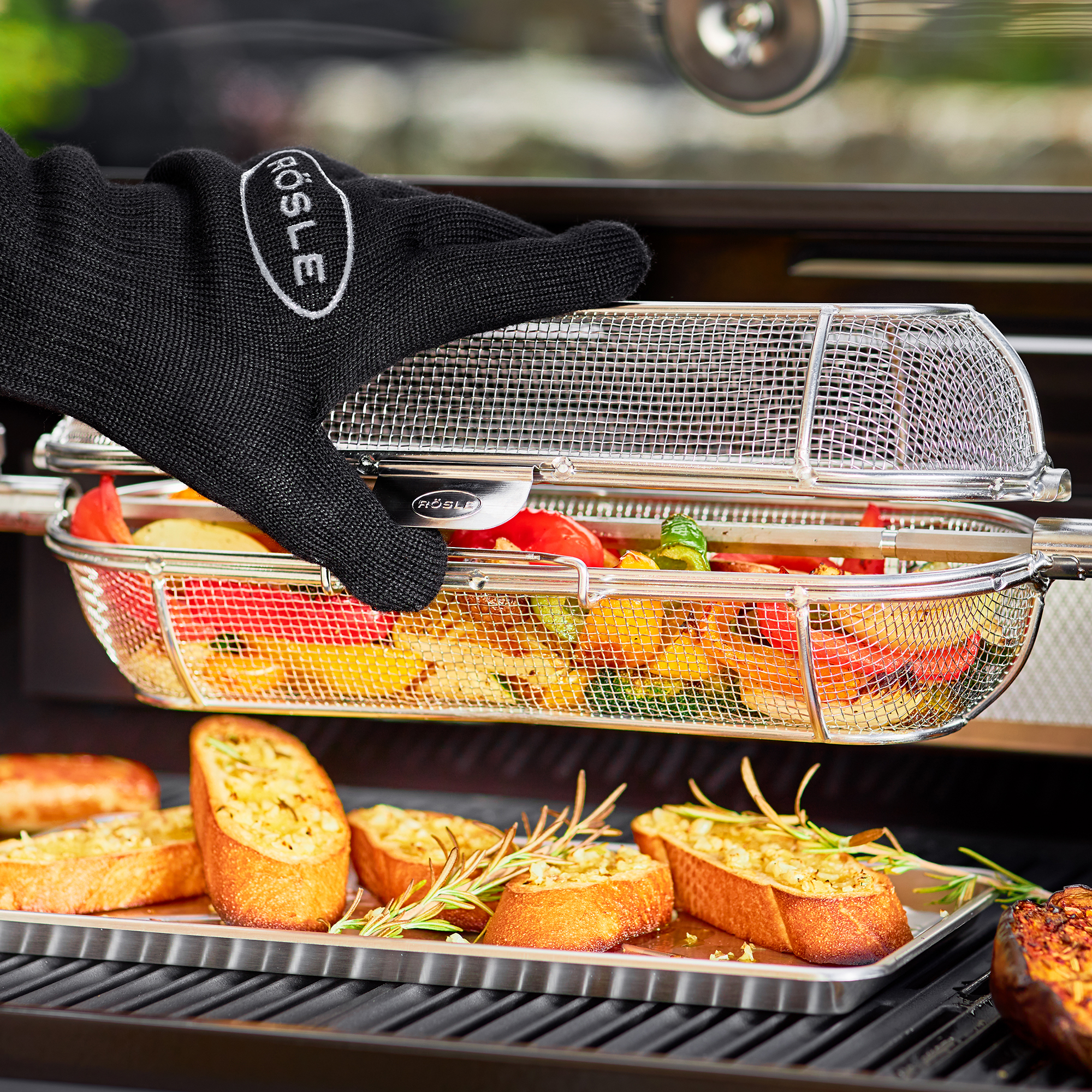 RÖSLE Drehspießkorb auf dem Grill Drehspießkorb mit Gemüse und Brot auf dem Grill, Hand mit Grillhandschuh greift nach dem Korb.
