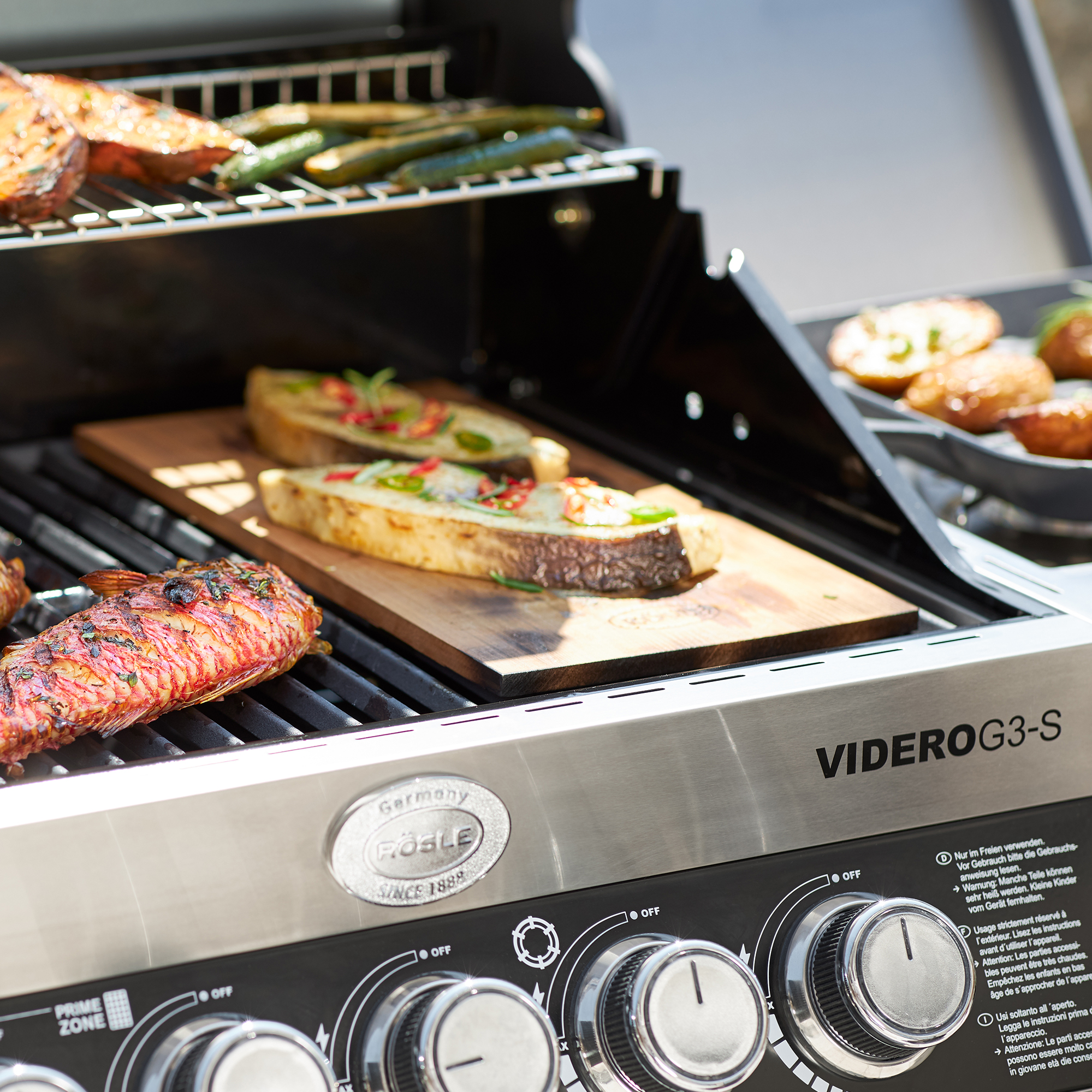 RÖSLE flavour plank alder on grill Flavoured alder plank in use on a grill, with various foods such as meat and vegetables.