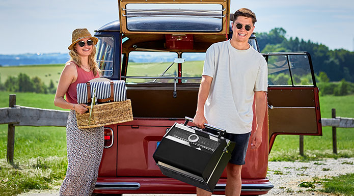 Elektrogrill E2-P Mann hält den mobilen Elektrogrill am Griff in einer Hand und Frau holt Picknick Sachen aus...