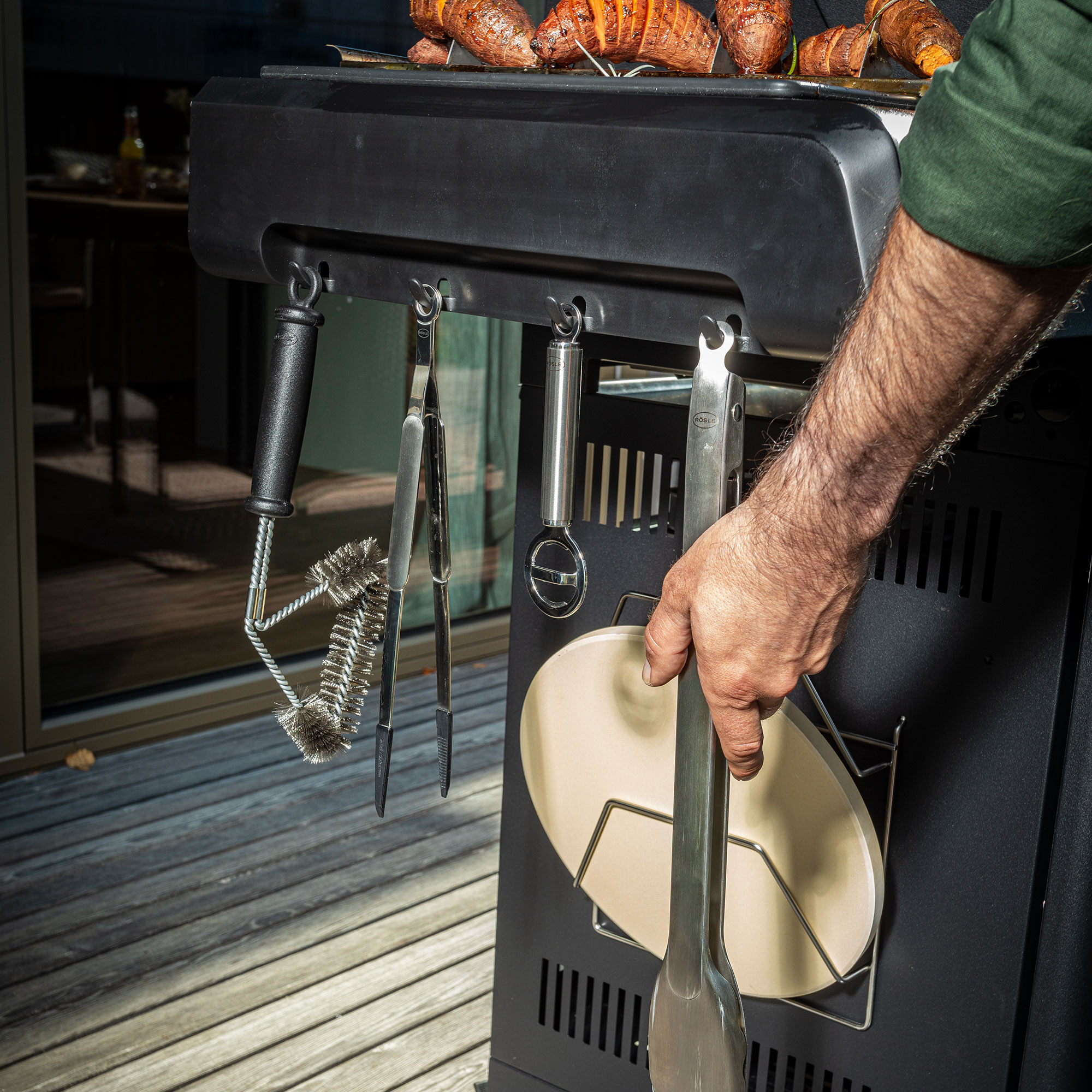 RÖSLE Grill mit Zubehör Grill mit Grillzubehör, darunter Zange, Wender und Grillbürste, in der Hand eines Mannes.